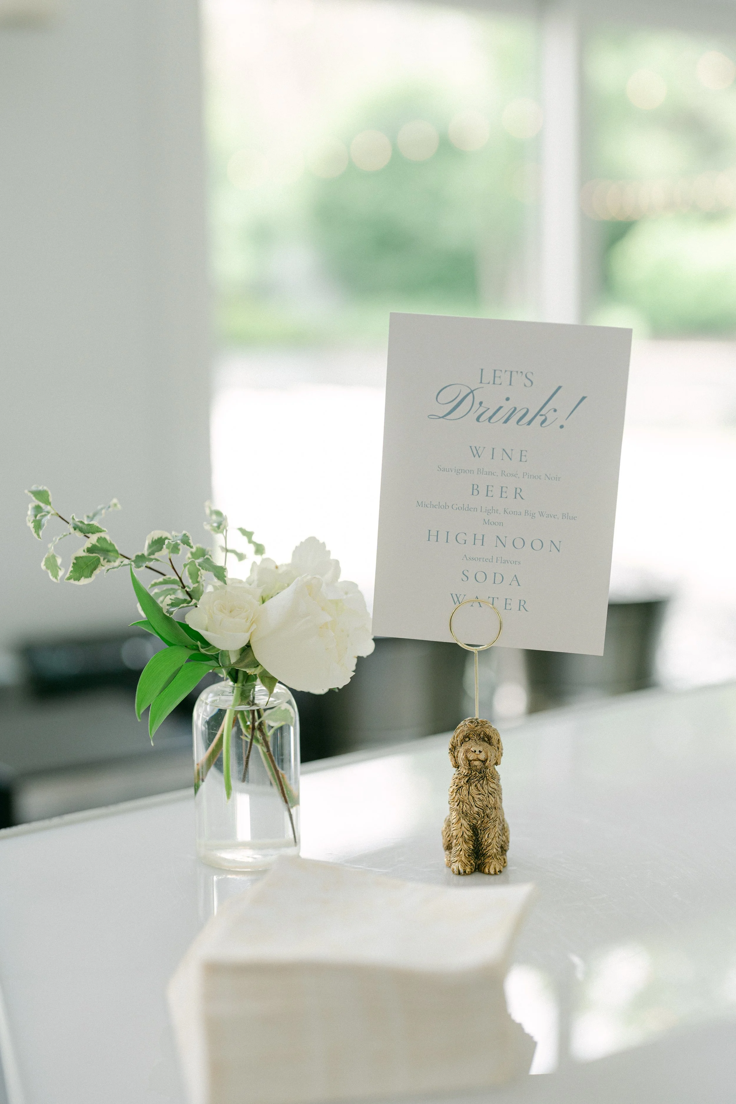 Decorative table centerpiece with a small glass vase holding white flowers and green foliage, a table card with menu items including wine, beer, high noon, soda, and water, and a small dog figurine. The background shows a bright, softly blurred windo