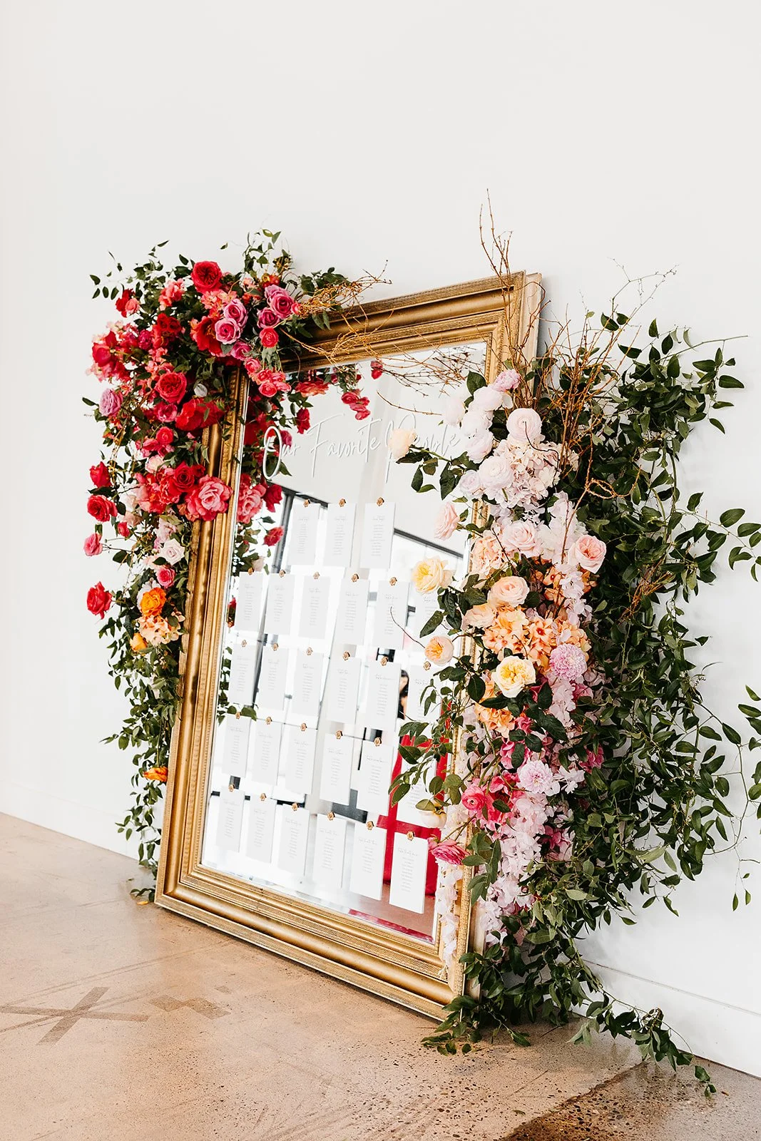 A decorative seating chart display with a large, ornate gold frame, decorated with pink, red, and white flowers and greenery, placed against a white wall.