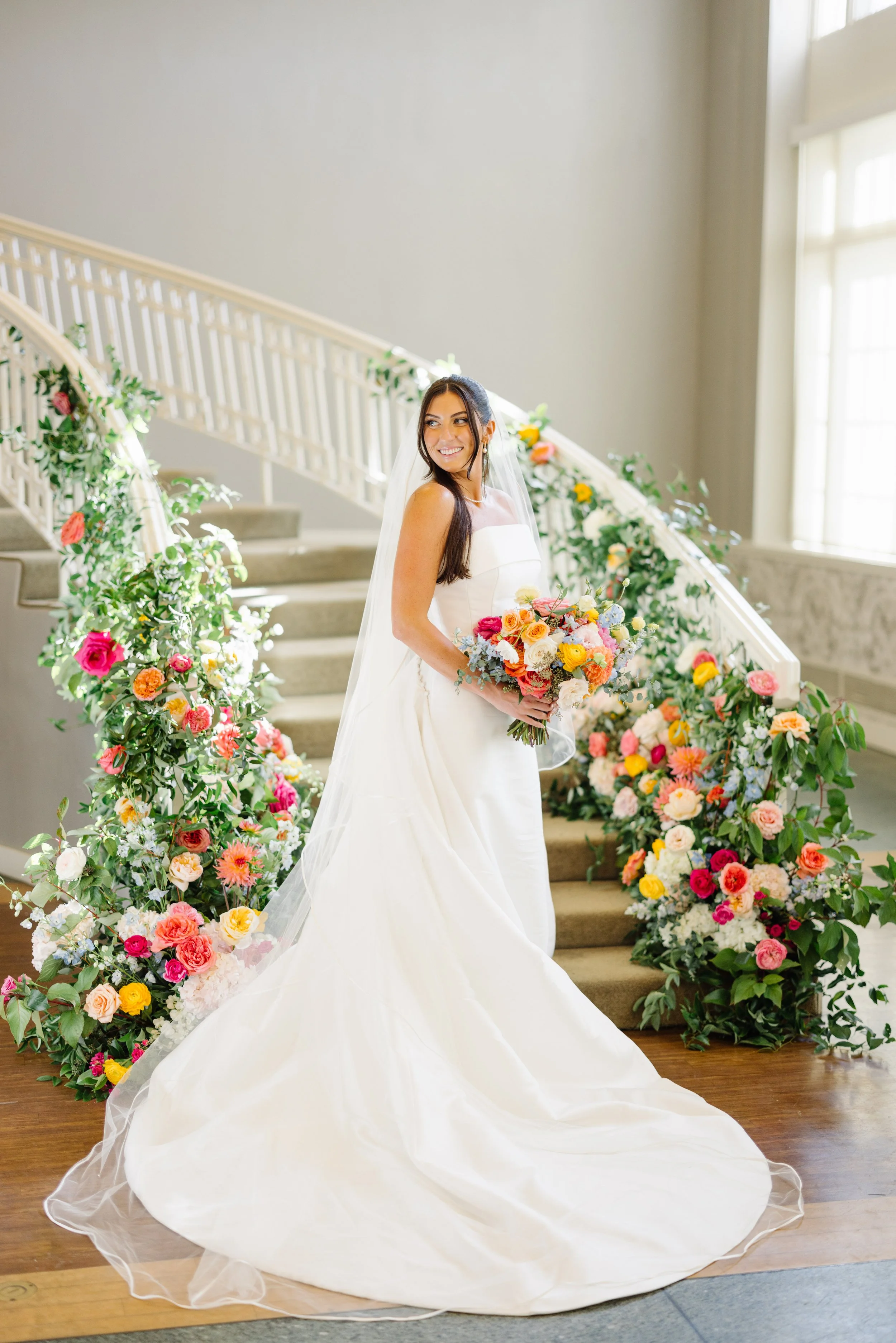 A bride in a white wedding gown holding a bouquet of colorful flowers, standing on a staircase decorated with flowers and greenery, indoors with large windows.