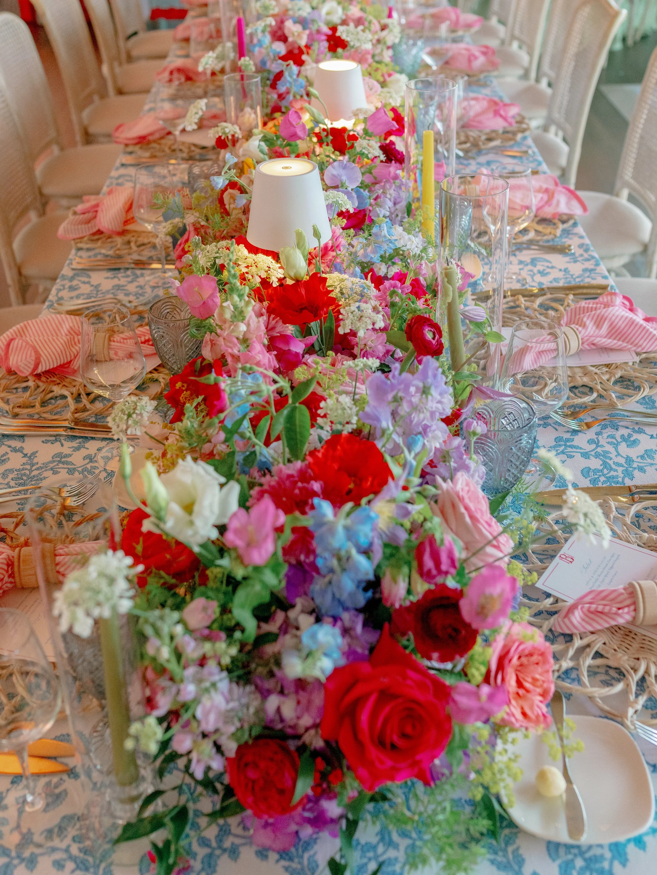 A long dining table decorated with a colorful floral centerpiece, candles, and place settings with pink napkins and glassware, set for a celebration or event.