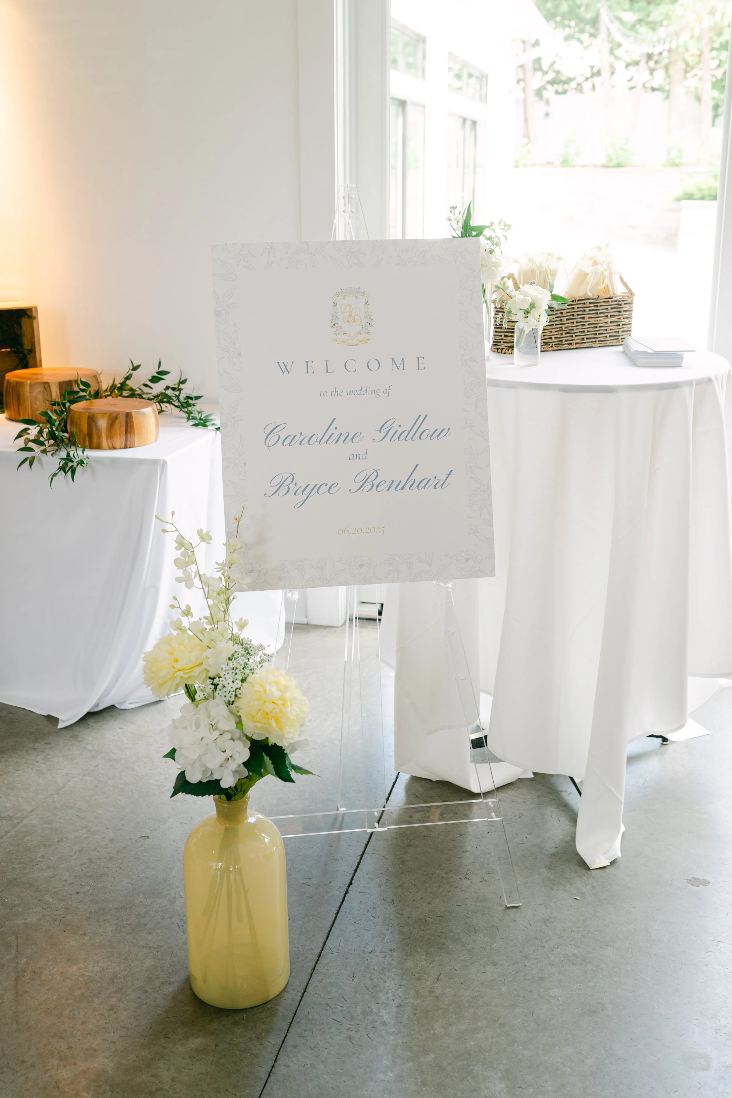 Wedding welcome sign with floral design, standing on a white easel, surrounded by white flowers and greenery, with tables and decorations in a bright, airy space.
