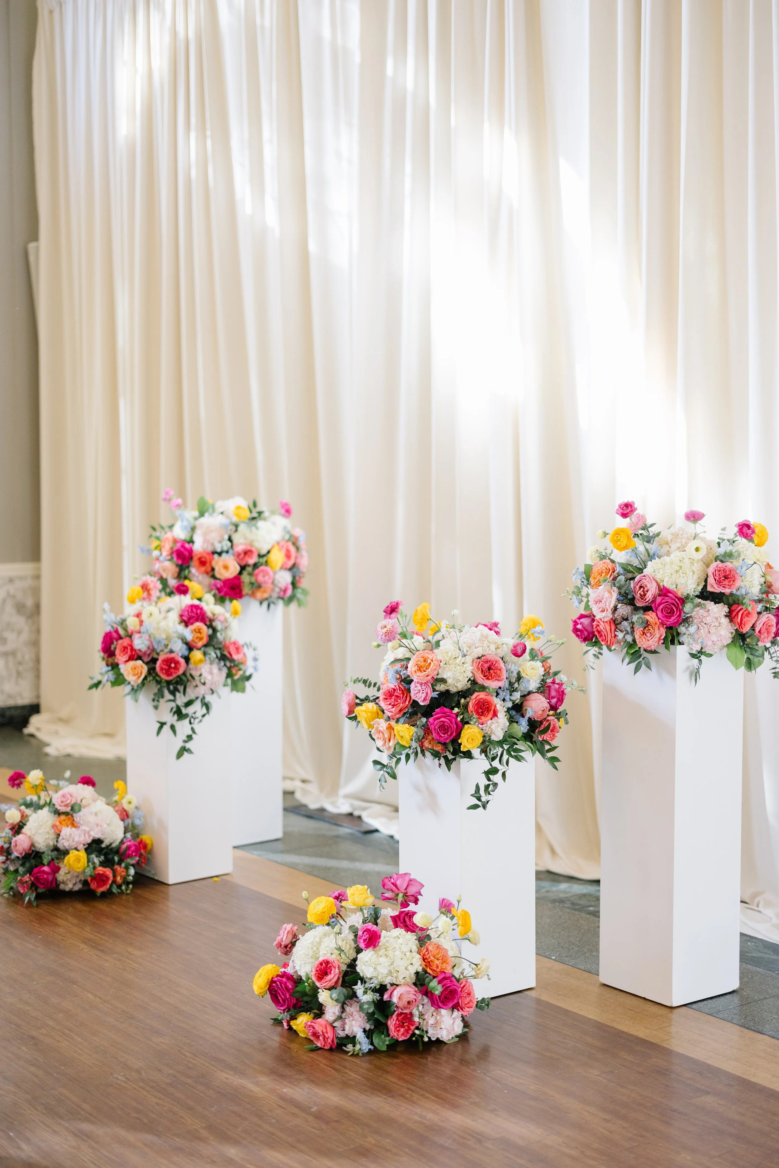 Elegant floral arrangements on white pedestals in front of cream-colored curtains, suitable for a wedding or formal event.