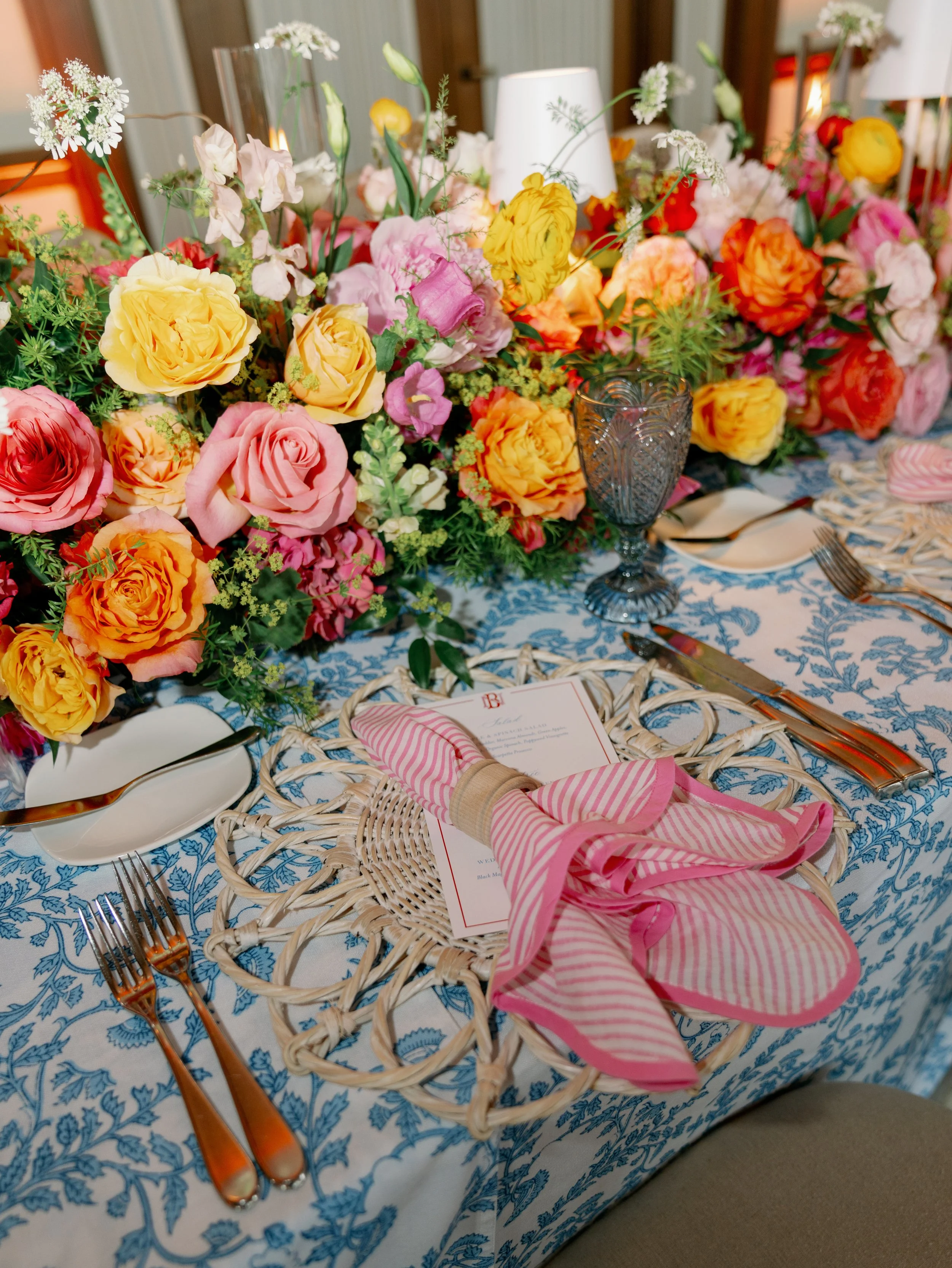 Table with floral centerpiece, blue patterned tablecloth, place settings including pink and white striped napkins, and amber-colored utensils.