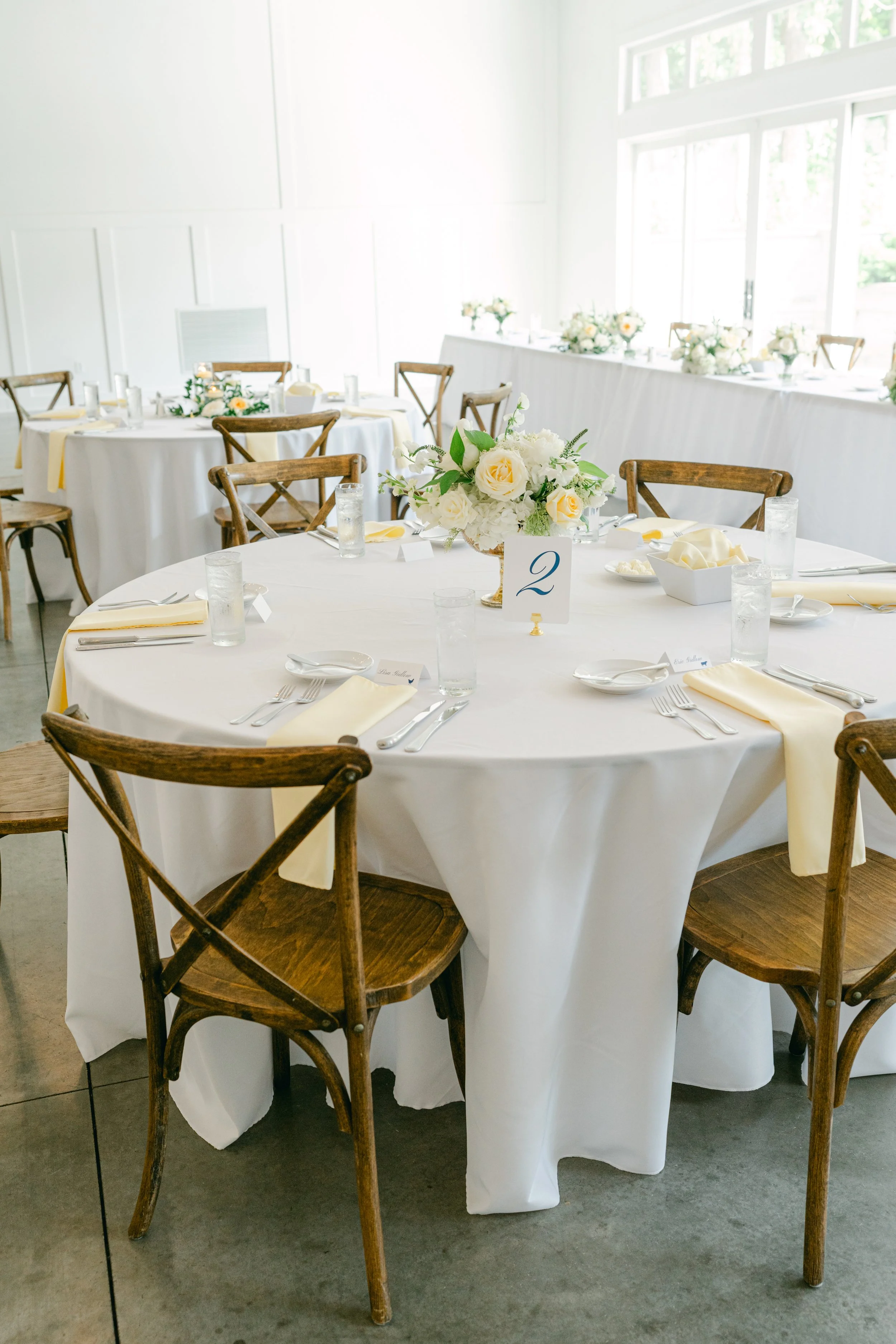 Round banquet table covered with white tablecloth, decorated with white and yellow floral centerpiece, surrounded by wooden chairs, set with napkins, silverware, and glasses, in a bright room with large windows and white walls.