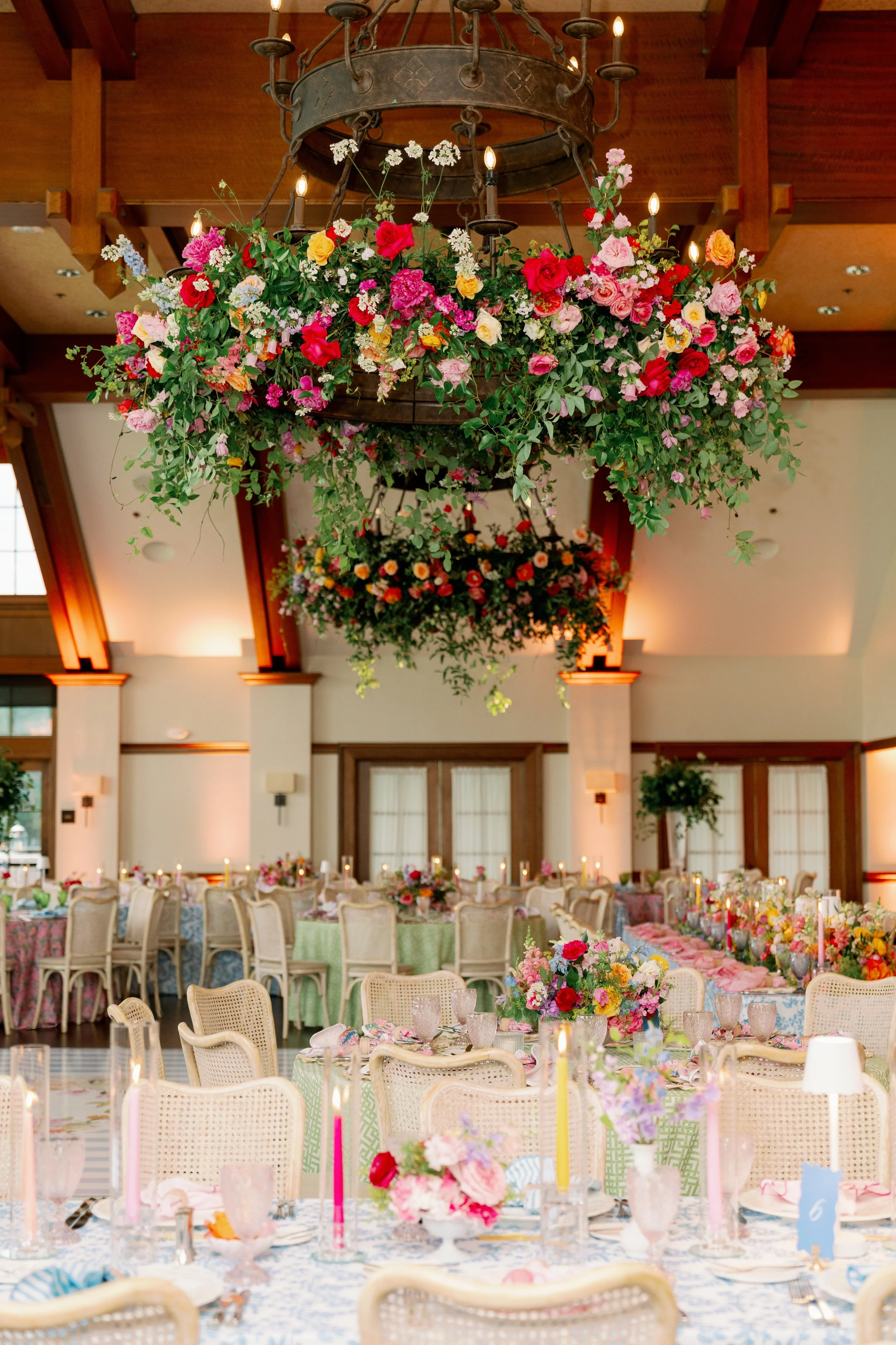 A large floral chandelier with roses and other flowers hanging from a rustic iron chandelier in a decorated banquet hall with table settings and chairs.