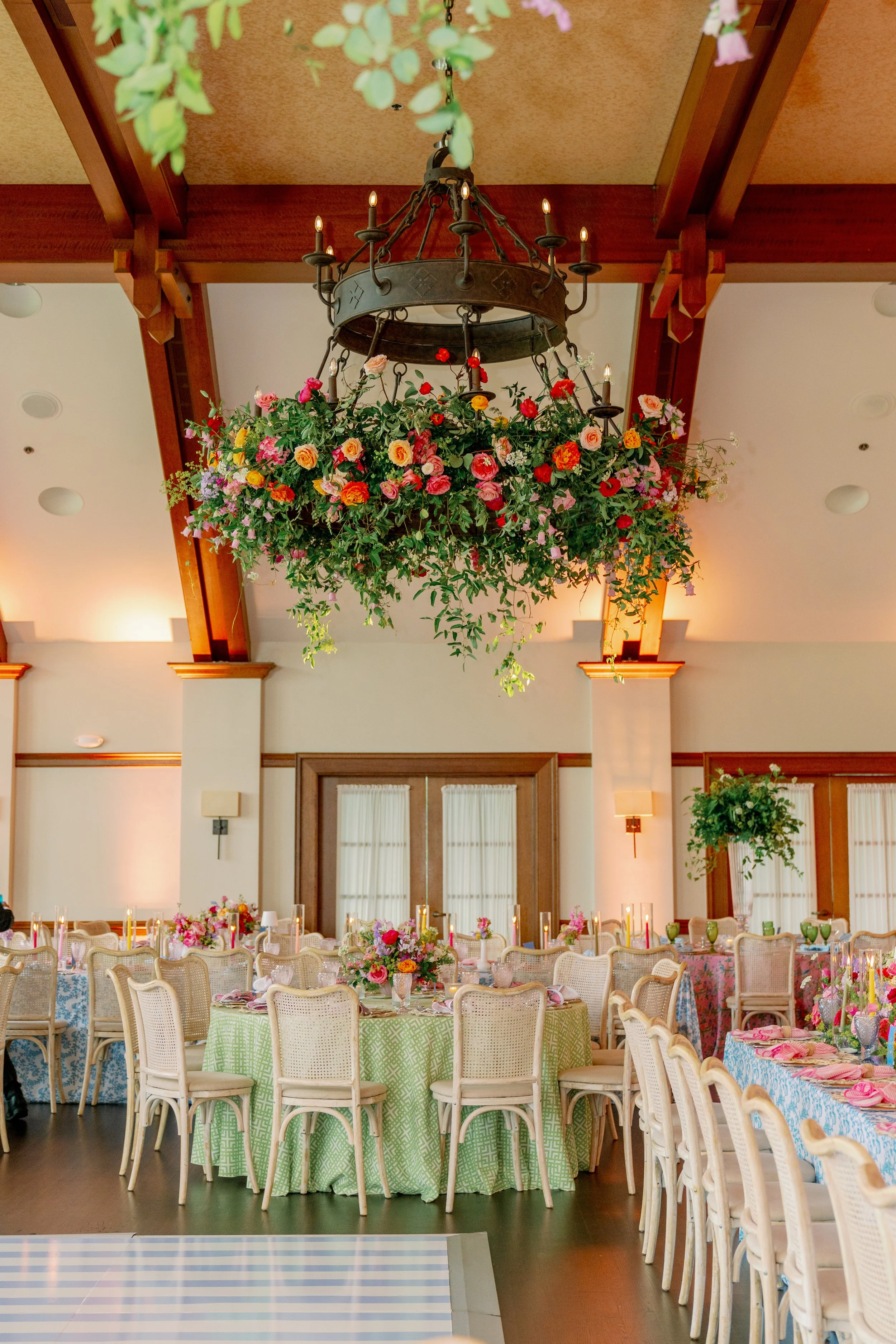 Decorated banquet hall with round tables covered in pastel green, pink, and blue tablecloths, each with floral centerpieces, surrounded by beige chairs. A large chandelier with candles hangs from the wooden ceiling with beams, above a floral arrangem