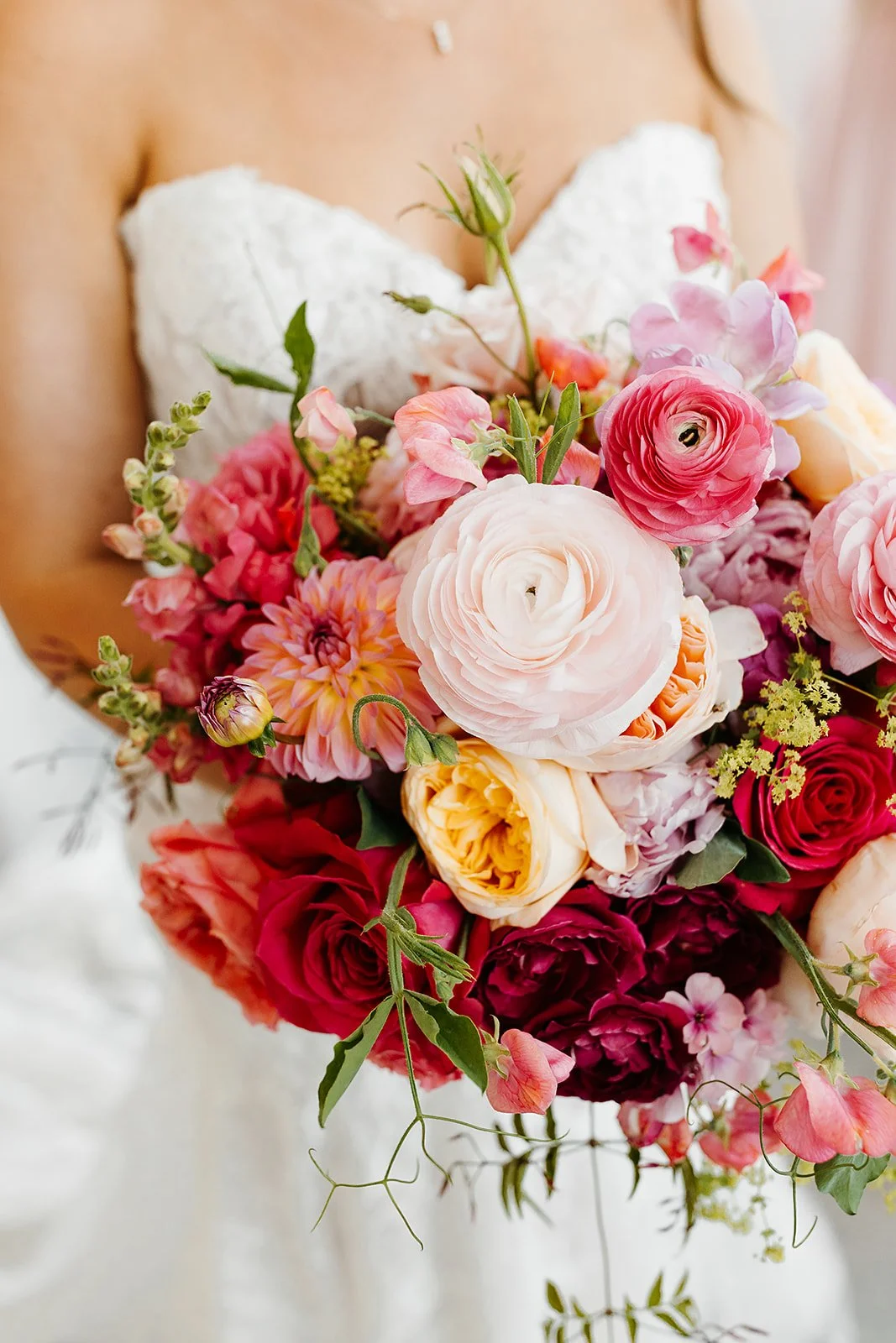 Close-up of a bridal bouquet with pink, white, and red flowers, held by a woman in a white dress with lace details.