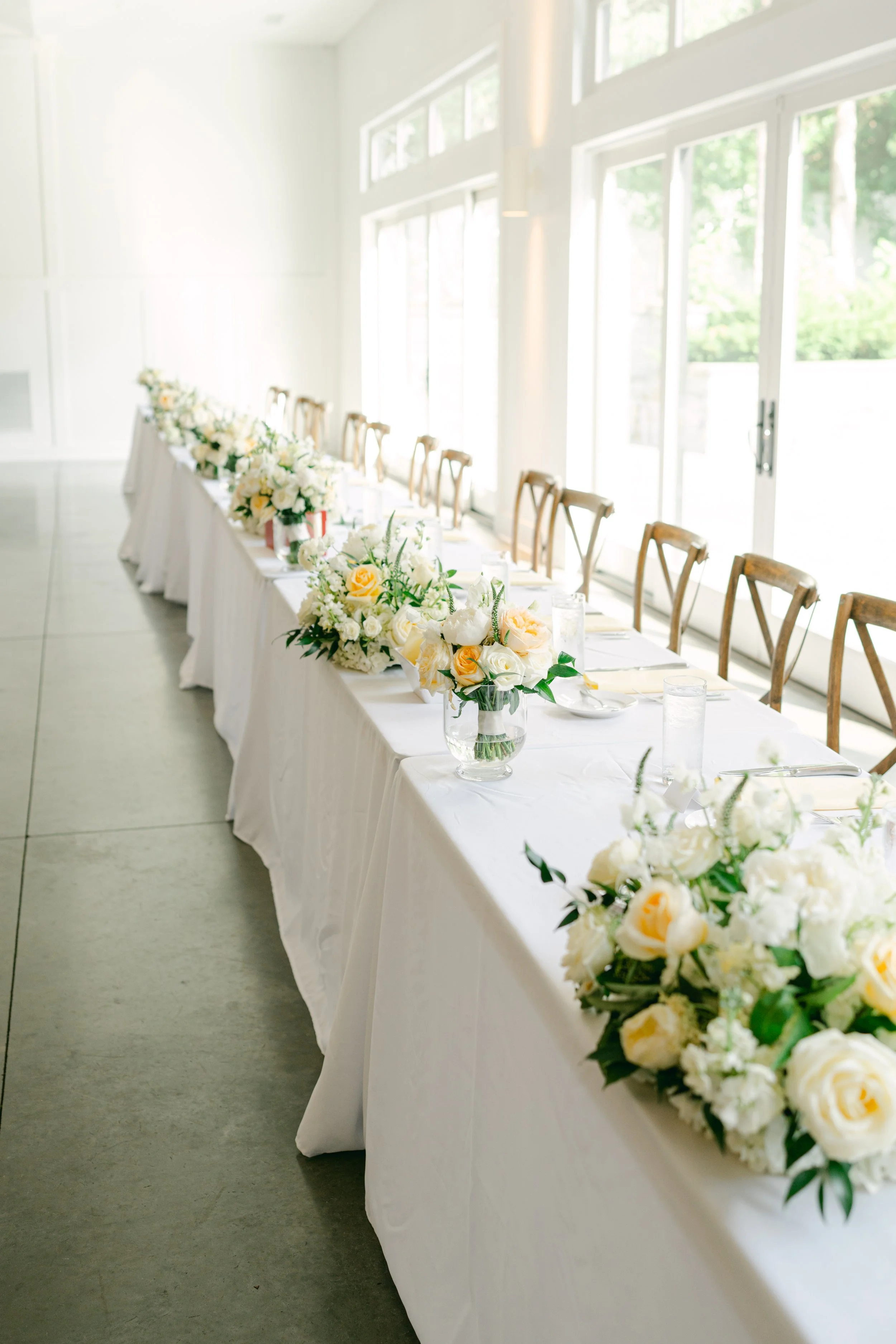 A long rectangular table draped with a white tablecloth, decorated with white and yellow floral arrangements, set with plates, glasses, and cutlery. The room is bright with large windows allowing natural light, and wooden chairs are aligned along the