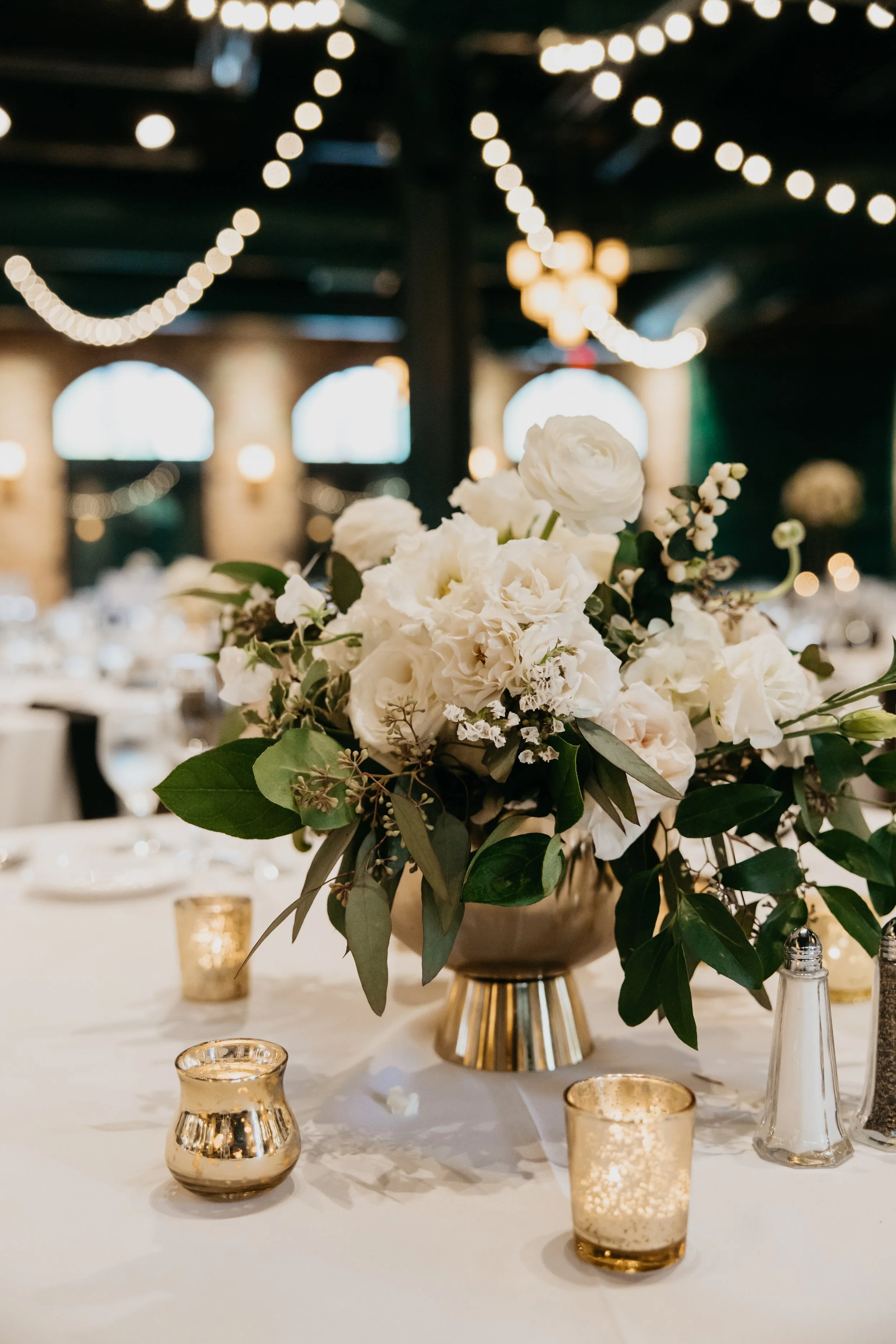 Close-up of a floral centerpiece with white flowers and green leaves in a gold vase, surrounded by small gold candle holders on a white tablecloth, in a dimly lit venue with string lights and arched windows in the background.