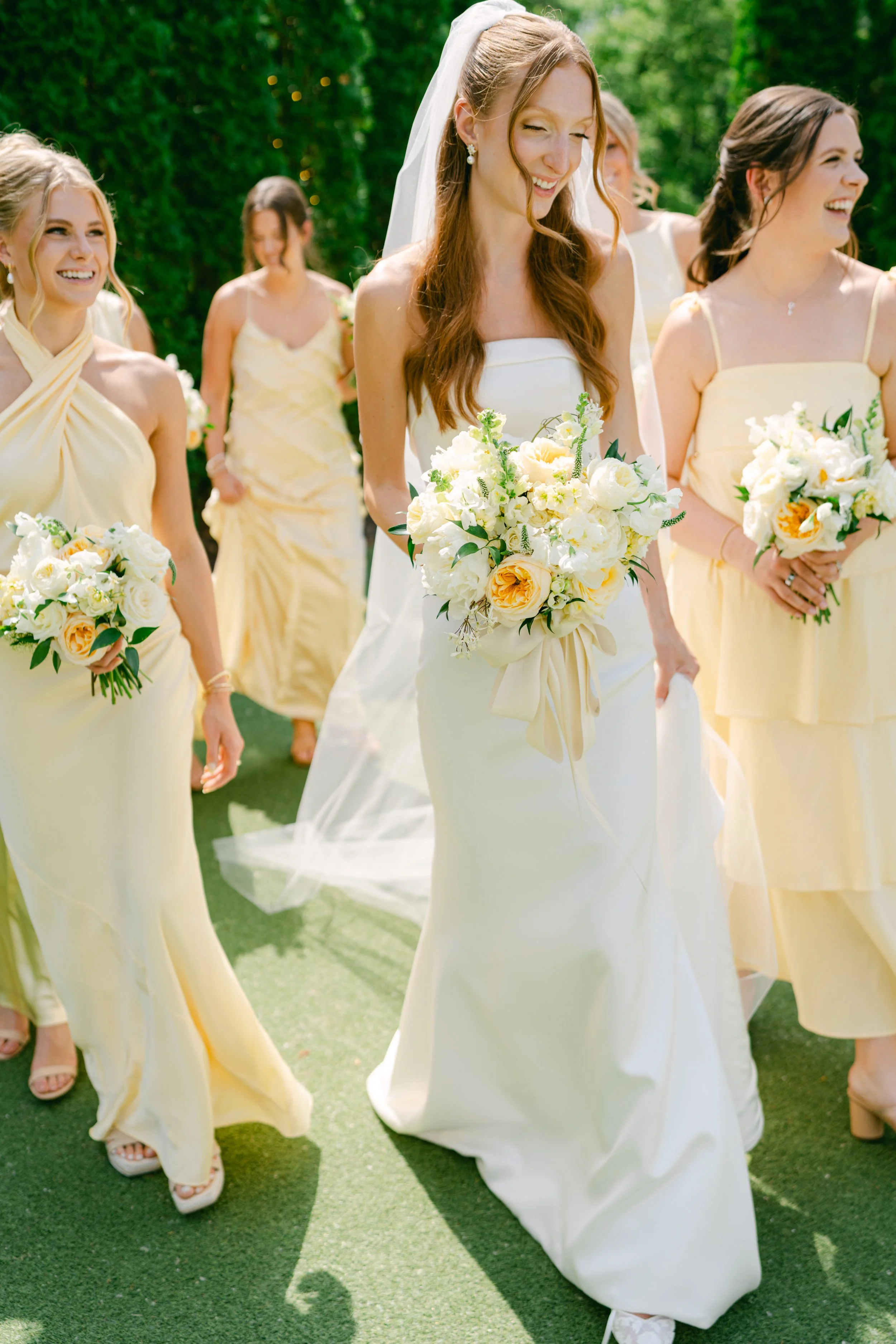 A bride in a white wedding gown holding a bouquet of white and yellow flowers, walking outdoors with bridesmaids dressed in pale yellow dresses, on a green grass area with a lush green background.