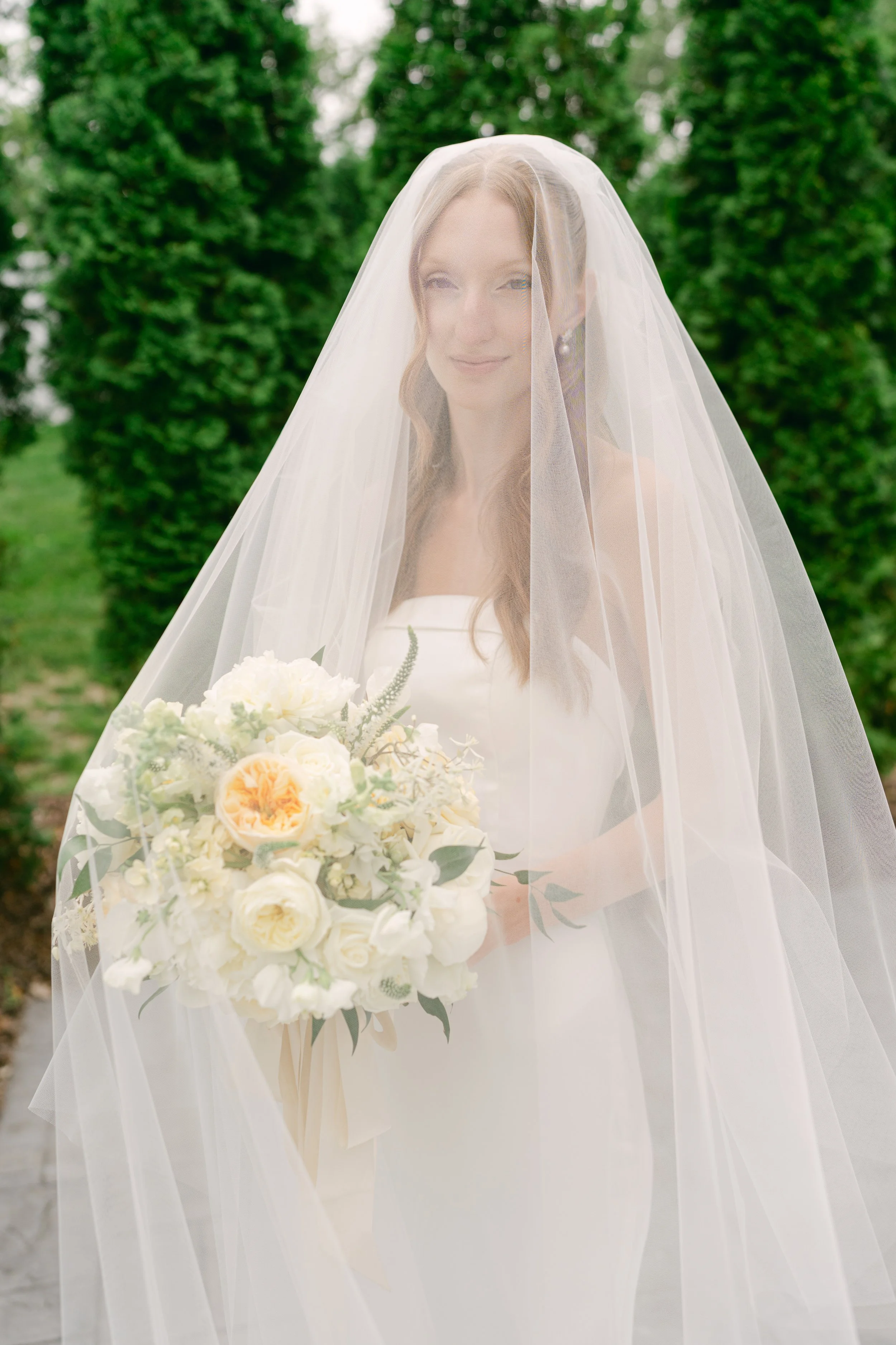 A bride stands outdoors with a radiant smile, wearing a white wedding dress and a sheer veil, holding a bouquet of cream-colored and peach flowers, with lush green trees in the background.