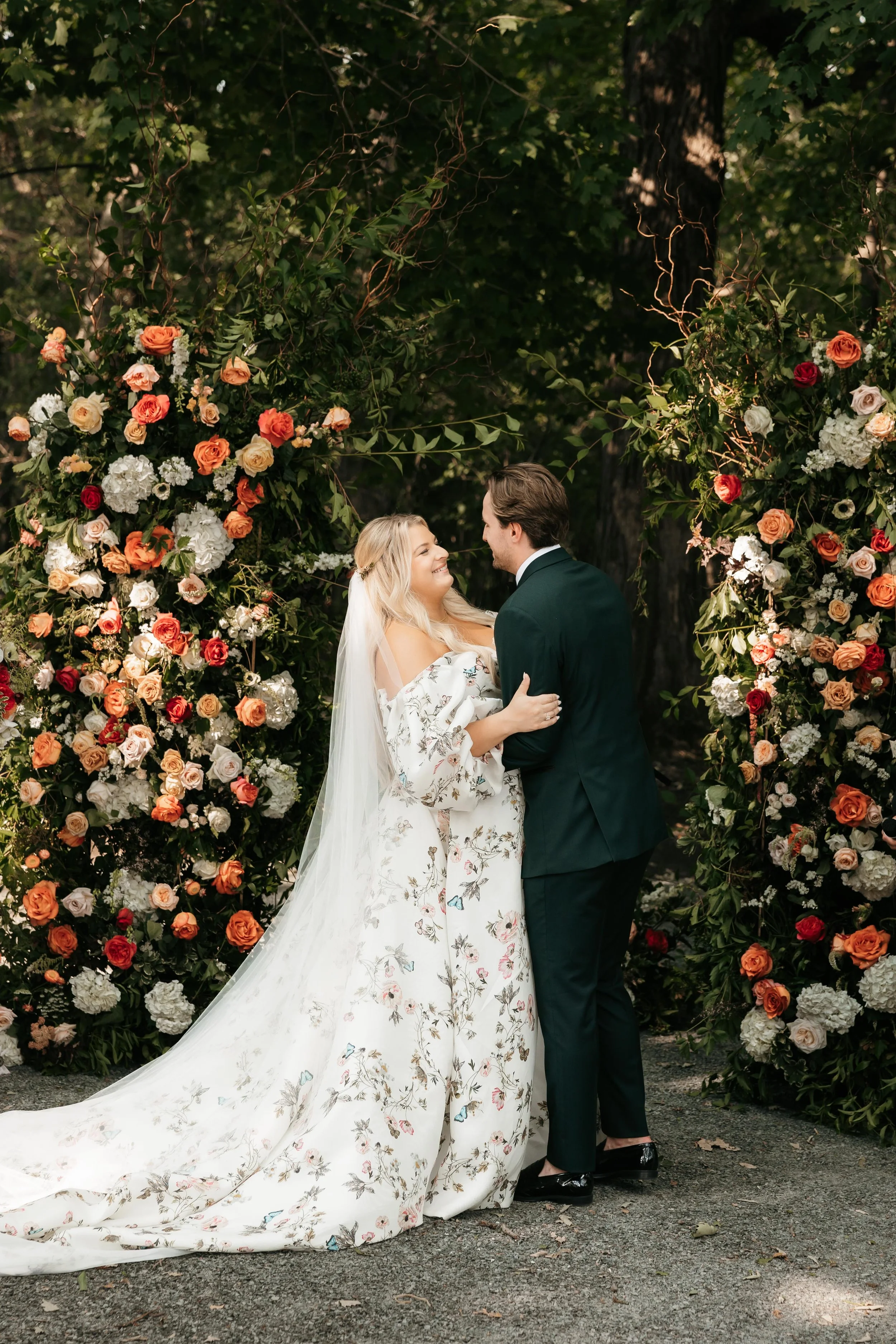 A bride and groom looking at each other and smiling, standing close together in front of a floral wedding arch with multicolored roses and white flowers, outdoors surrounded by trees.