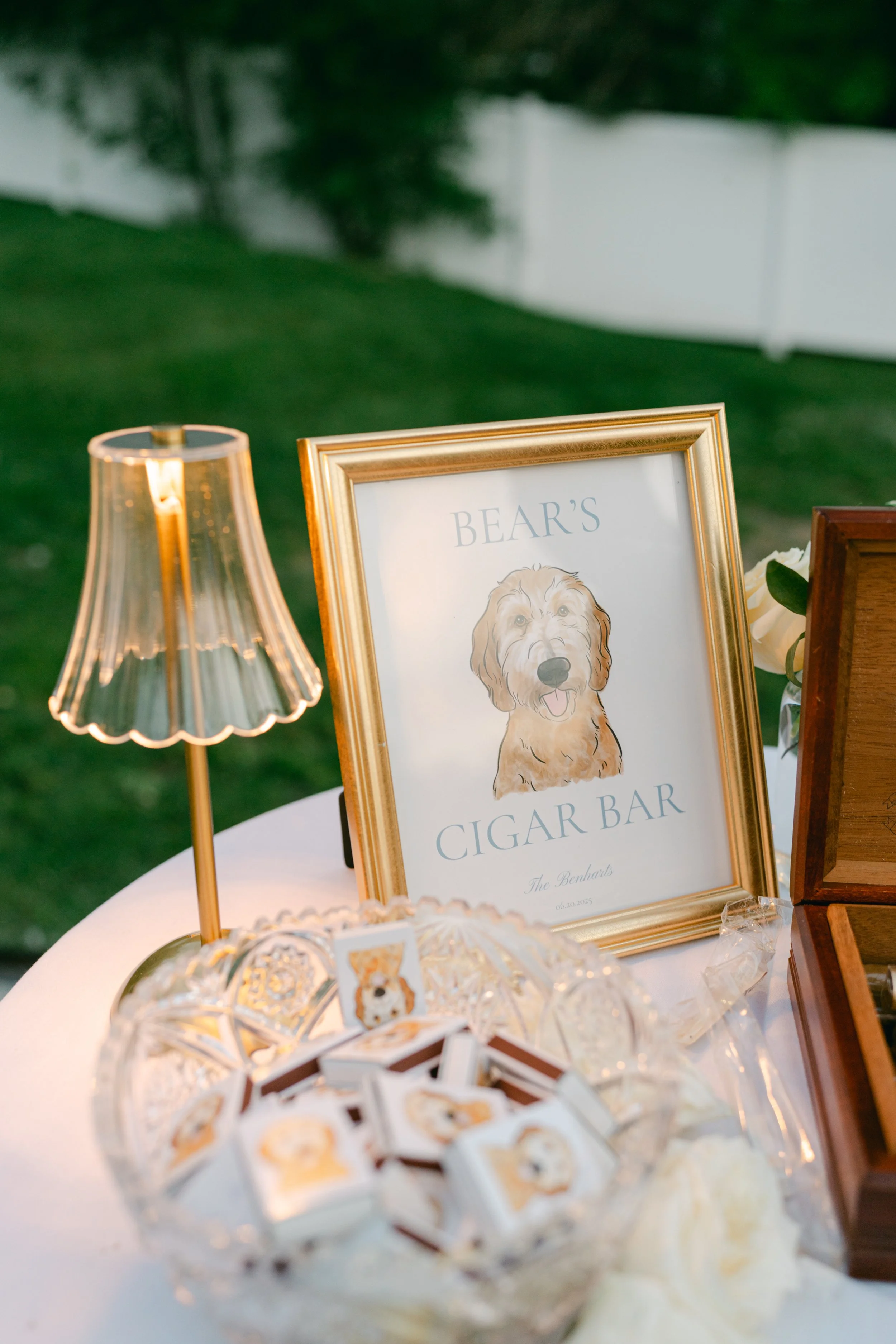 A framed sign for a cigar bar with a picture of a golden retriever dog, beside a small lamp and cupcake-shaped decorations on a round table.
