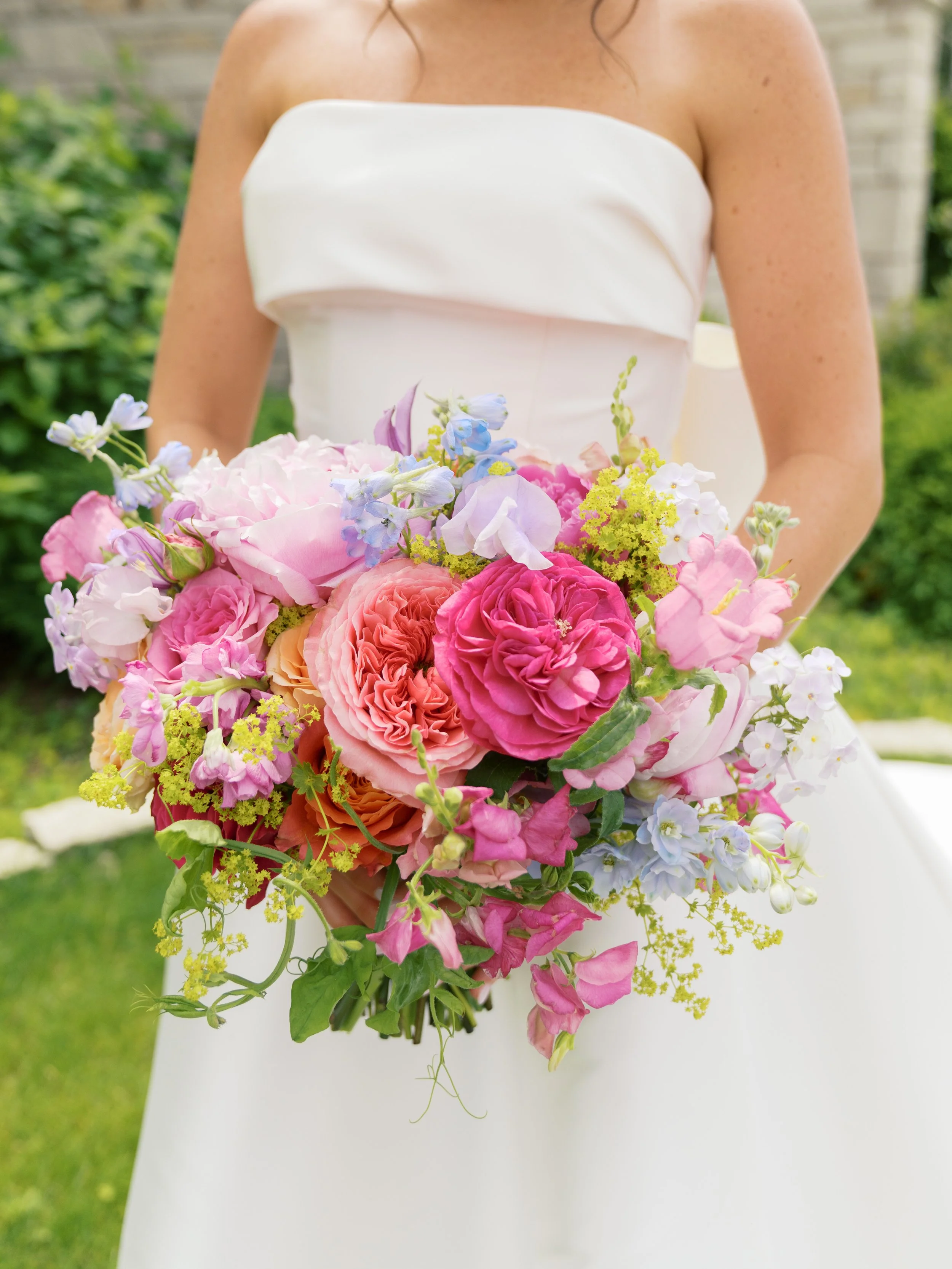 A woman in a white strapless dress holding a large colorful bouquet of flowers outdoors.