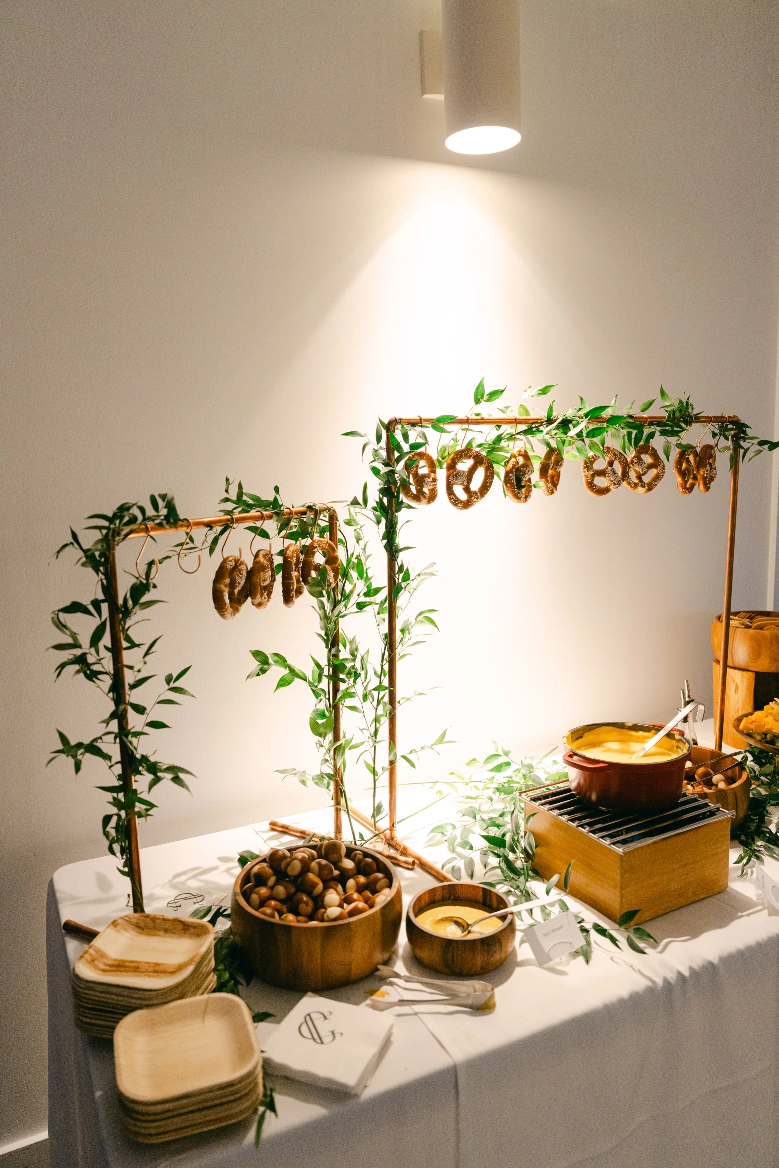 A buffet table with wooden bowls filled with nuts, plates, and napkins, decorated with green leaves. There are two clothing racks with pretzels hanging on them and a pot of cheese or dip on a small grill. The setup is in a well-lit room with a white 