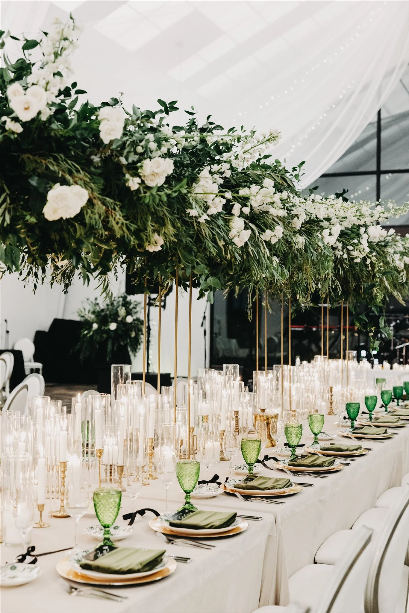 Long banquet table decorated for a formal event with a lush green and white floral arrangement overhead, surrounded by chairs with white covers, and set with elegant glassware, plates, and napkins.