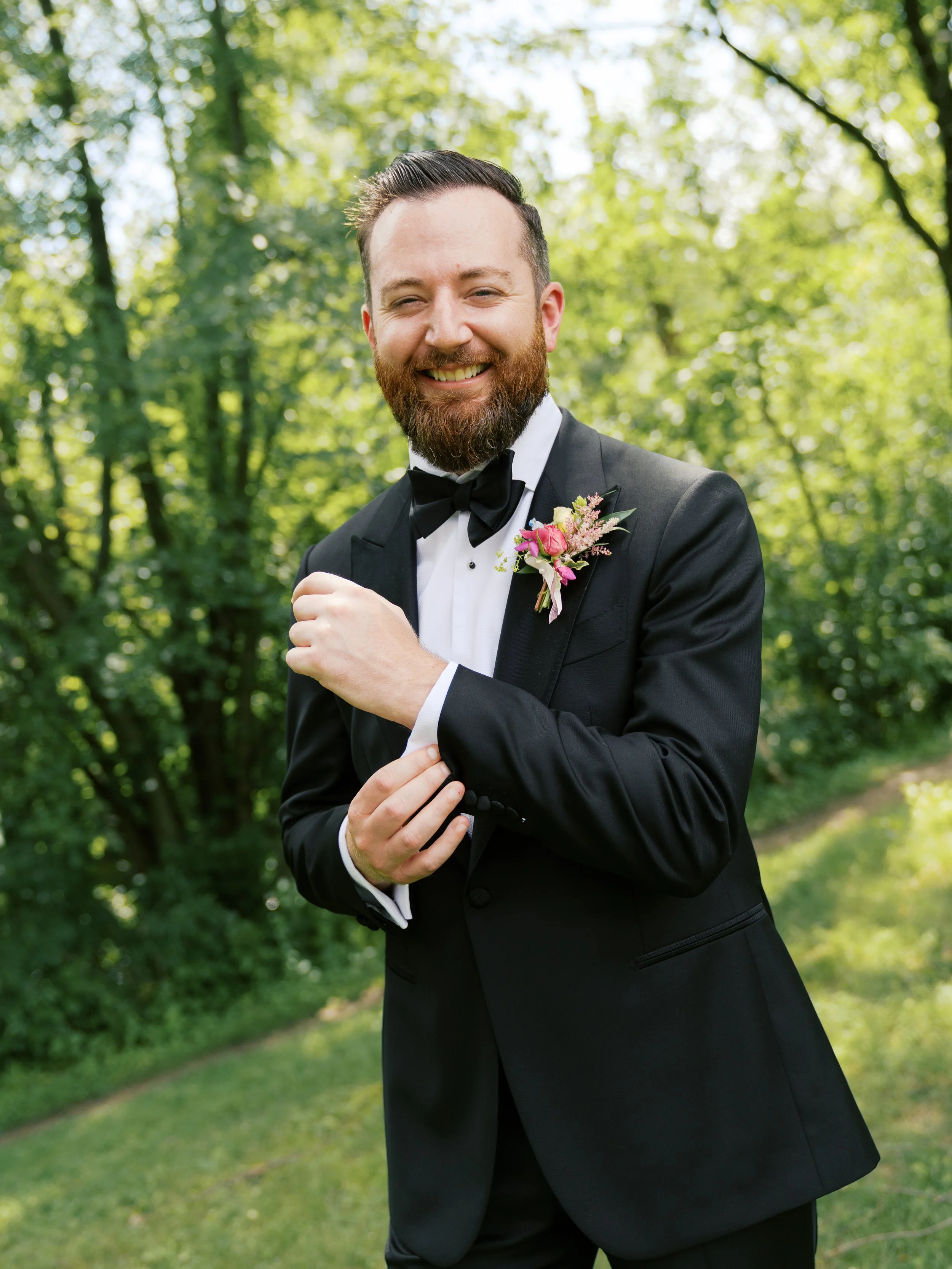 A man in a tuxedo with a boutonnière, standing outdoors among green trees, smiling and adjusting his jacket.