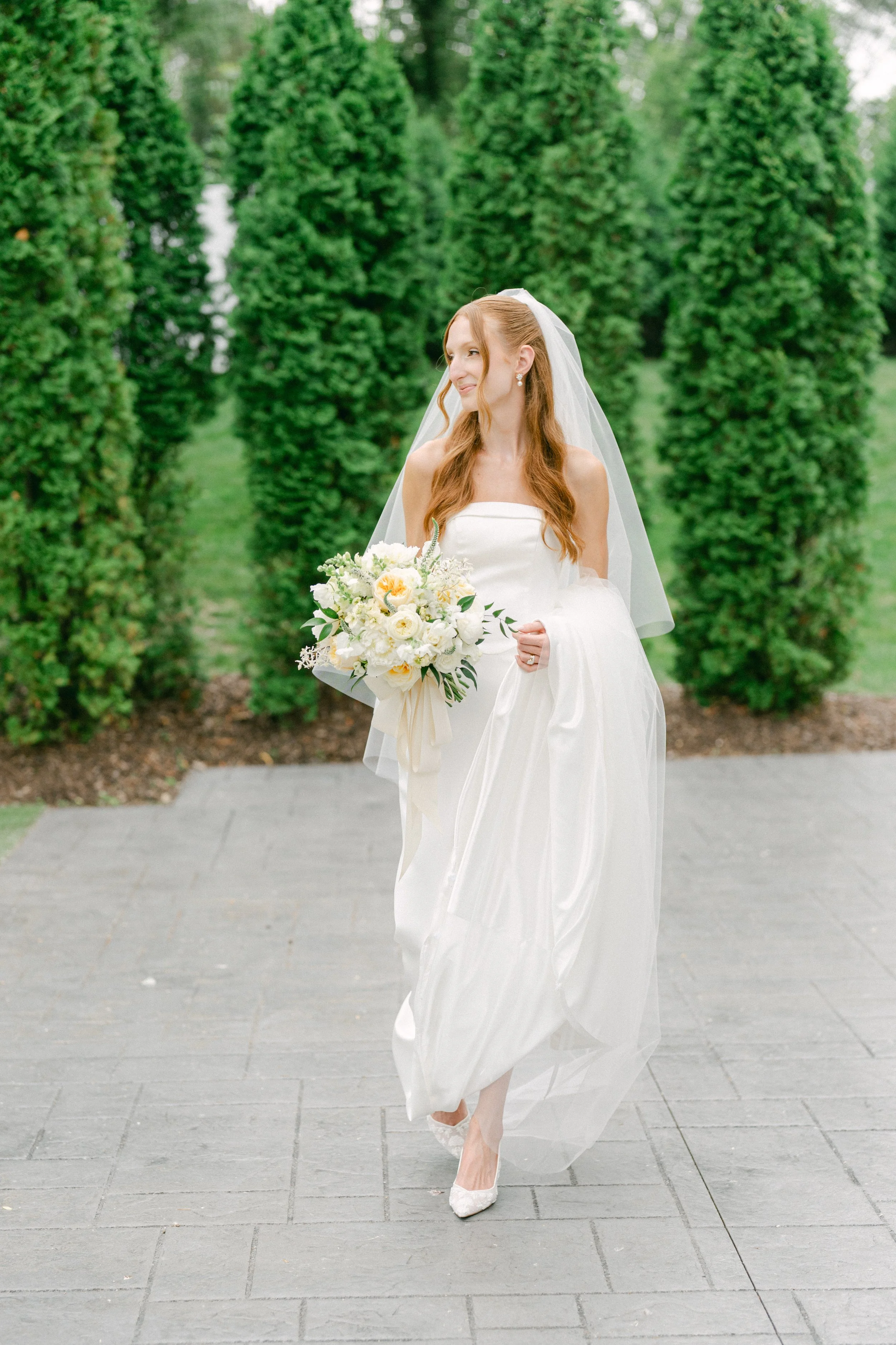 A woman in a wedding dress holding a bouquet of white flowers, standing outdoors on a paved walkway with tall, green, manicured bushes in the background.