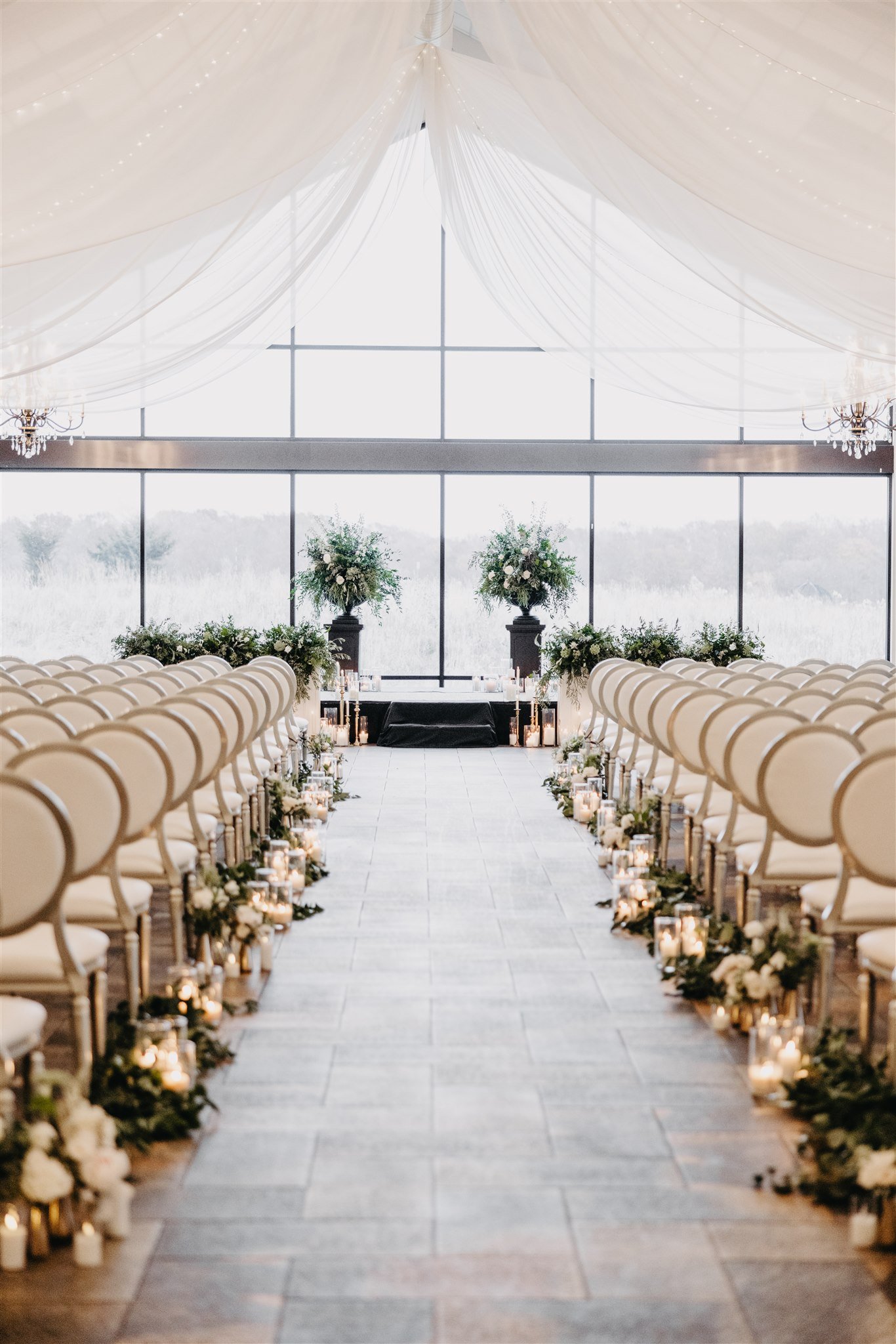 Elegant indoor wedding ceremony setup with rows of white chairs, flower arrangements on stands, and candles lining the aisle, under a draped ceiling with string lights and large windows in the background.