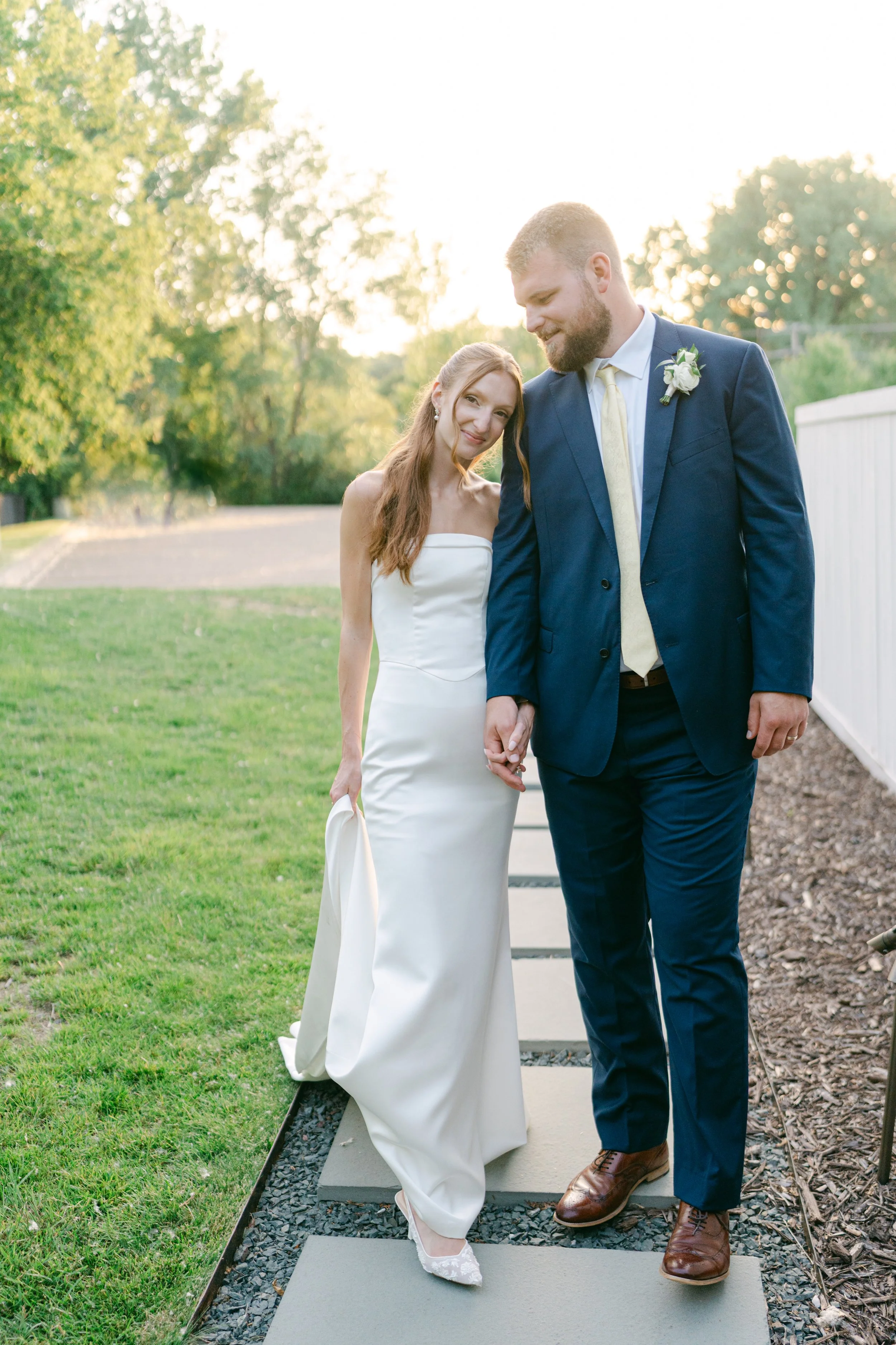 A bride and groom holding hands outside during sunset, with trees and a white fence in the background.