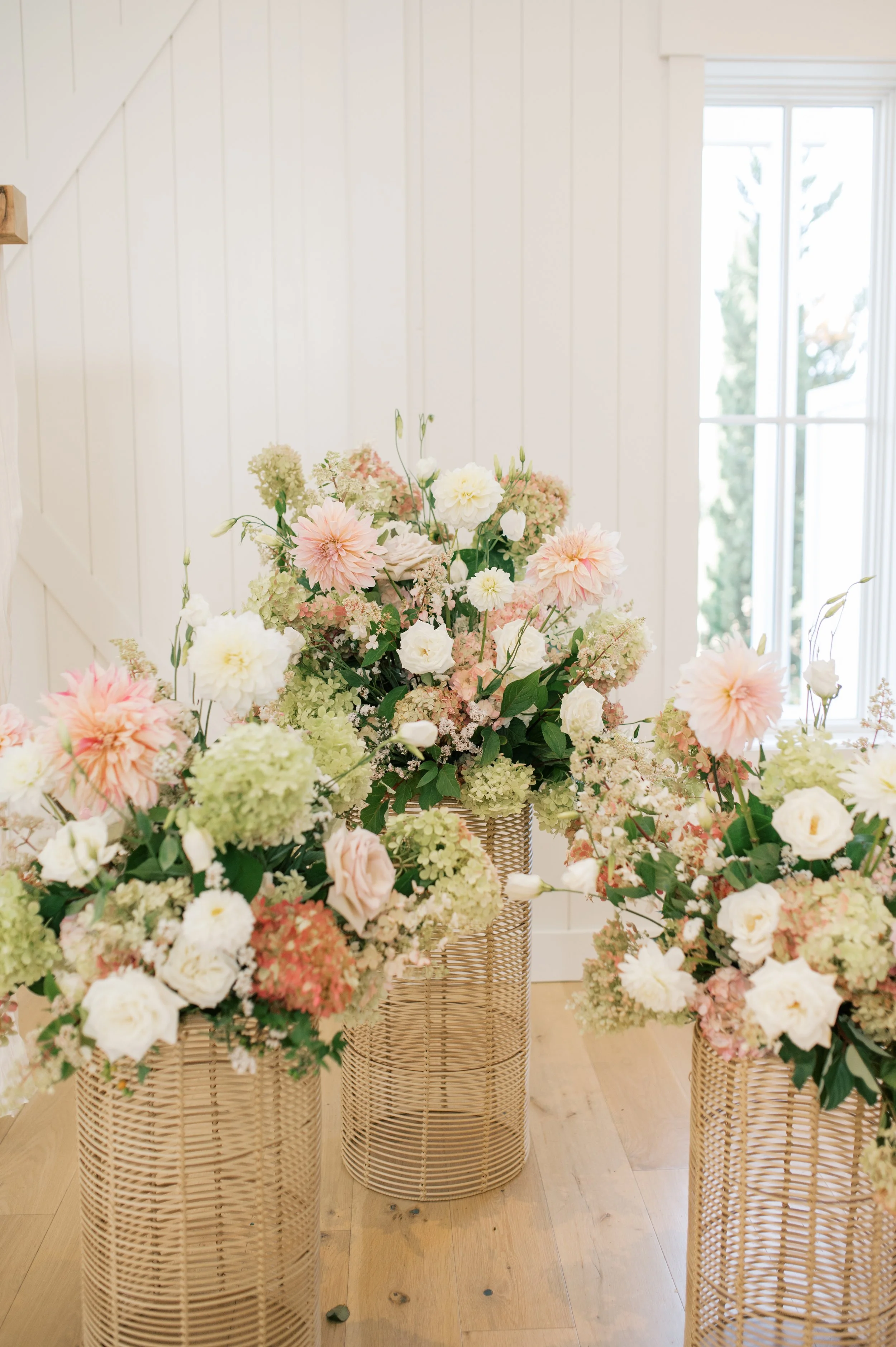 A display of tall wicker baskets filled with pastel-colored flowers, including pink, white, and green hydrangeas, set in a well-lit room with white paneled walls and large windows.