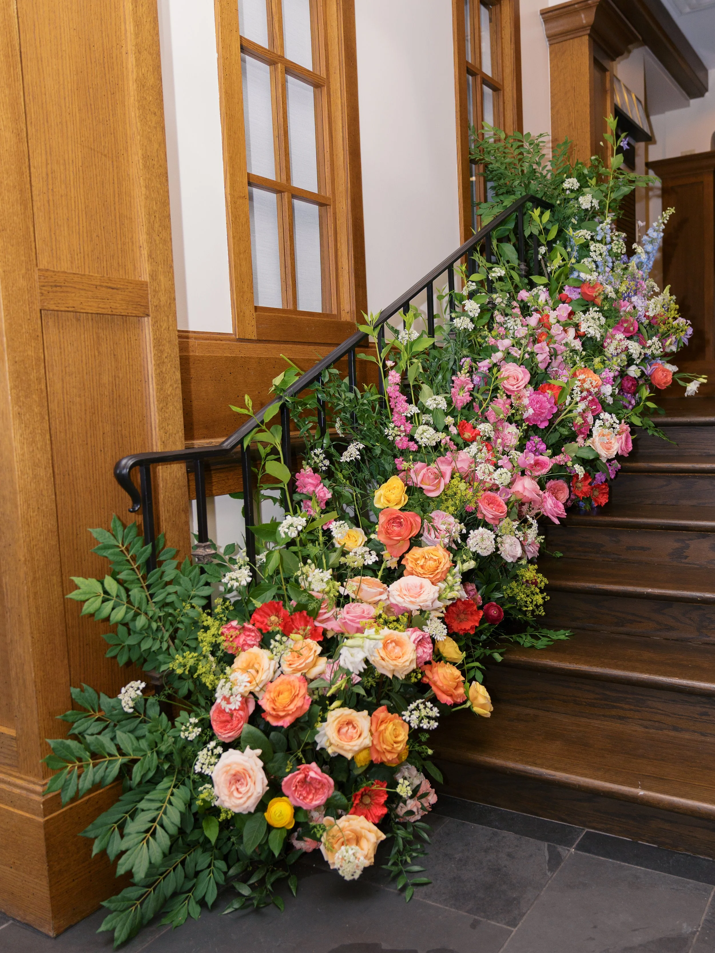 A large, colorful floral arrangement placed at the base of a staircase with wooden paneling and a black metal railing.