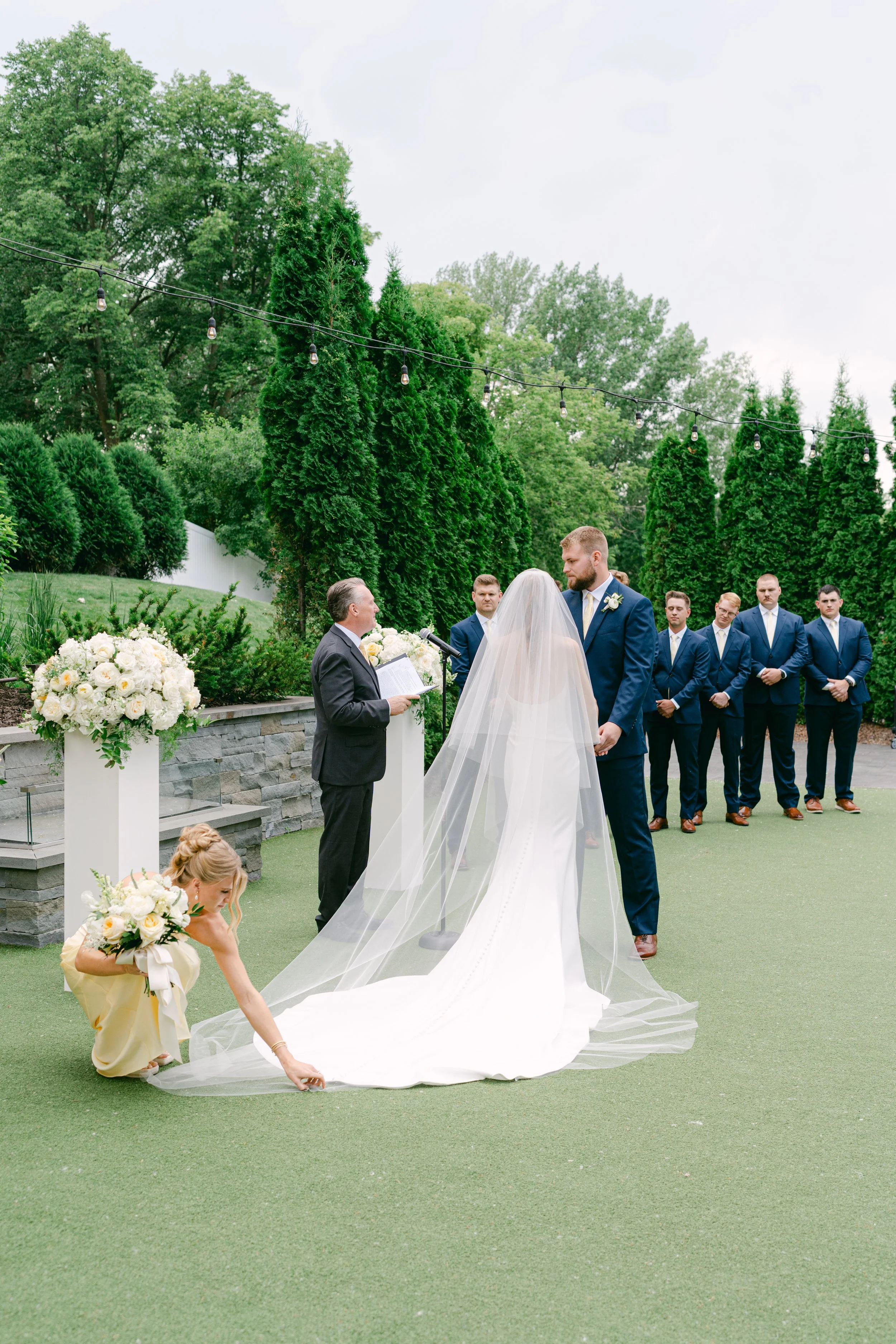 A wedding ceremony taking place outdoors with a bride and groom standing before an officiant, surrounded by groomsmen and a flower girl, with lush green trees and string lights overhead.