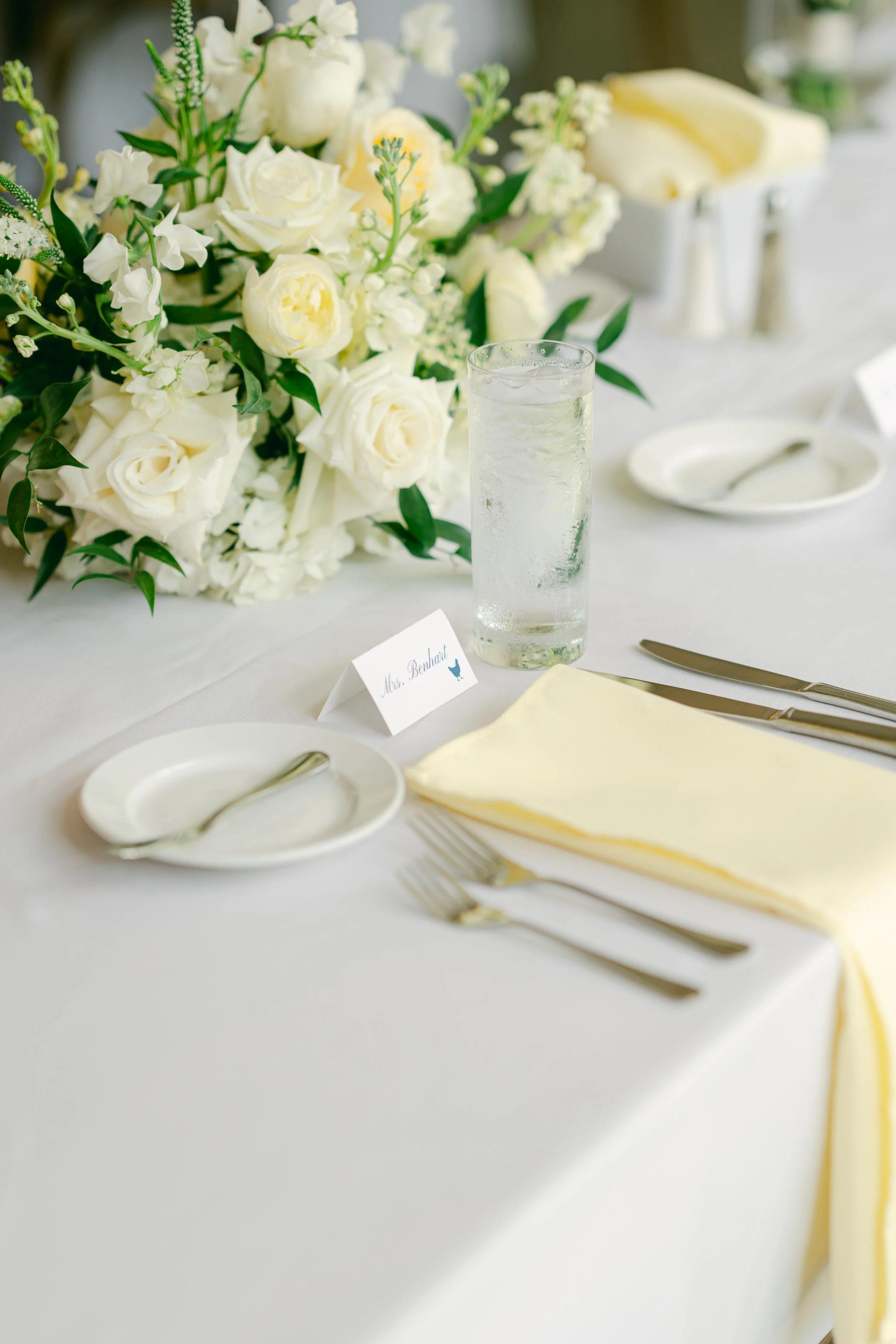 A wedding reception table setup with a large white floral centerpiece, a glass of water with ice, a white plate with a spoon, and a folded yellow napkin, with a name card reading 'Mrs. Behrart' on the table.