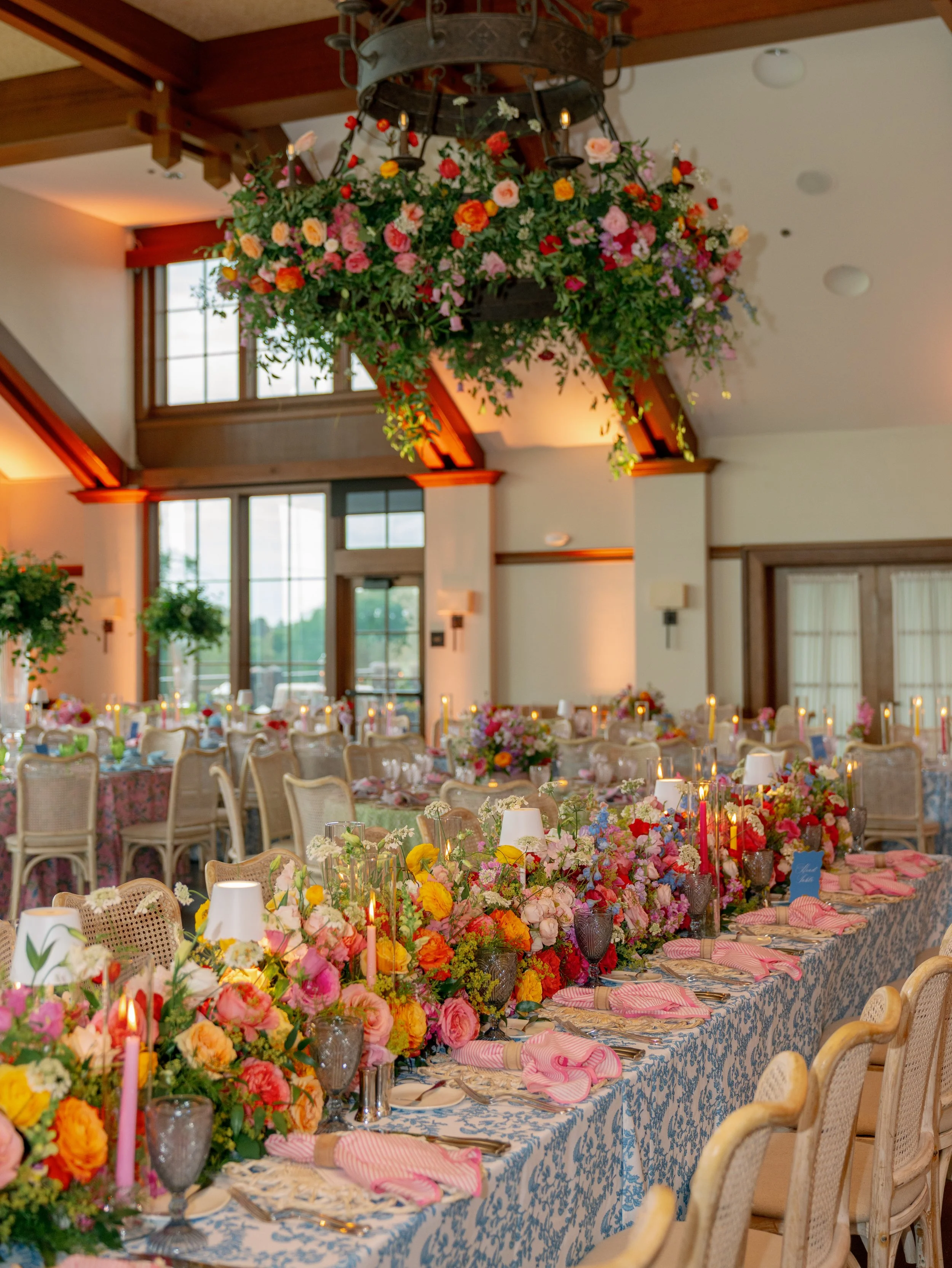Elegant banquet table decorated with large colorful floral arrangements, candles, and pink napkins in a beautifully decorated room.