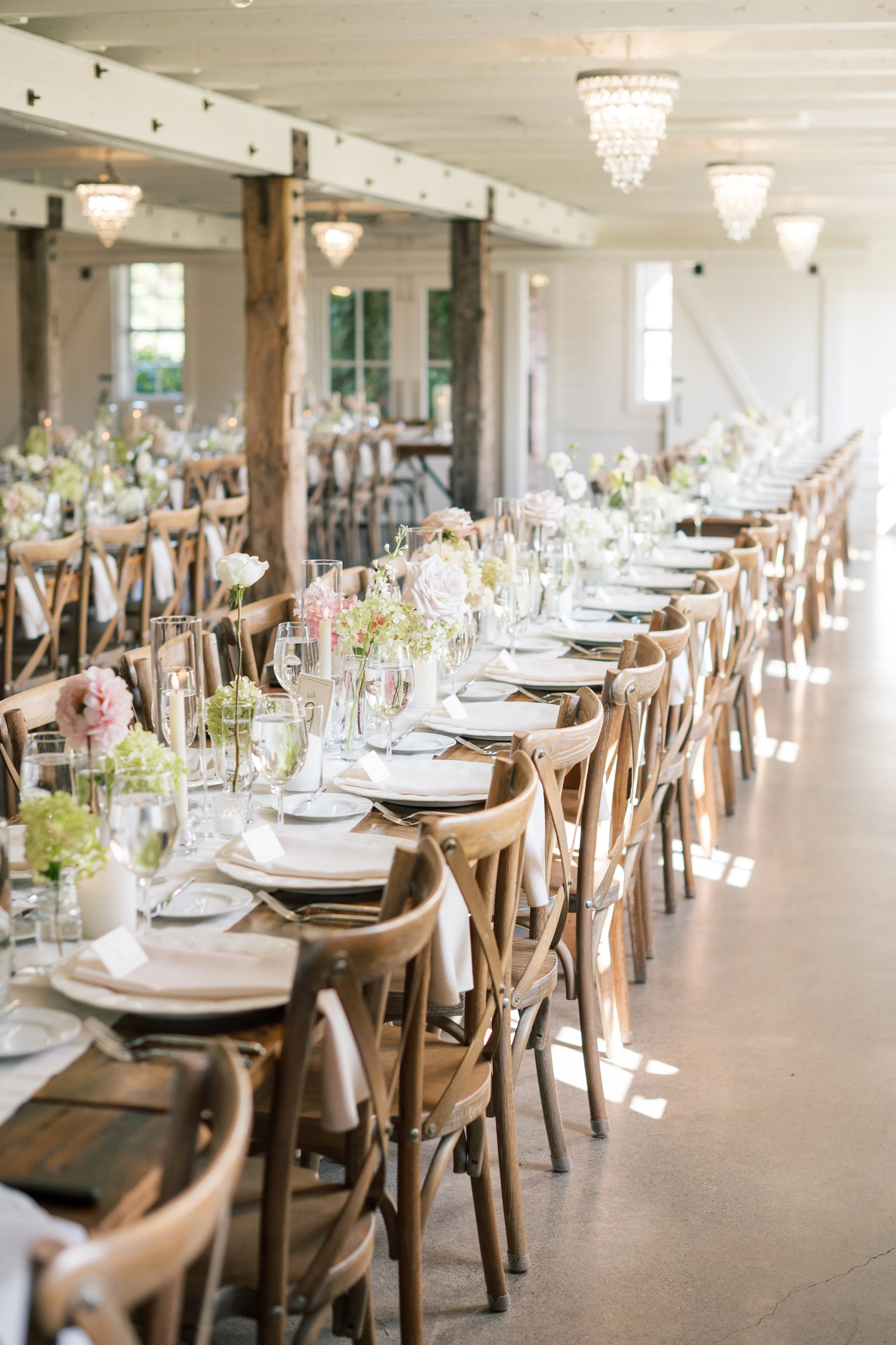 Long dining table in rustic style banquet hall beautifully decorated with flower arrangements, candles, glassware, and napkins, set up for a wedding or special event, with wooden chairs, chandeliers, and natural light.