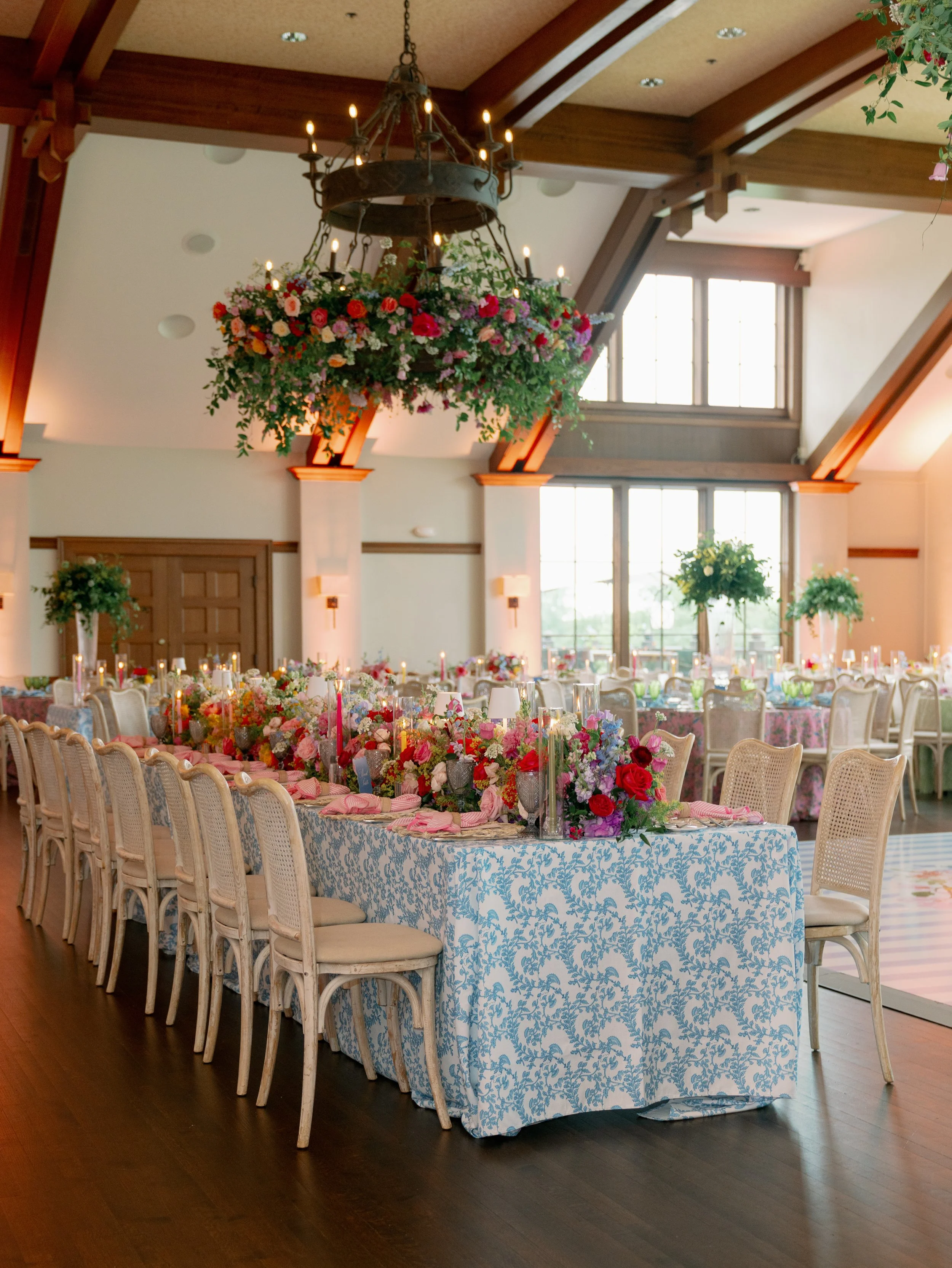 A large banquet table decorated with a blue and white damask tablecloth, floral centerpieces, and candles, set up in a spacious room with wooden beams, large windows, and a chandelier with candles hanging from the ceiling.