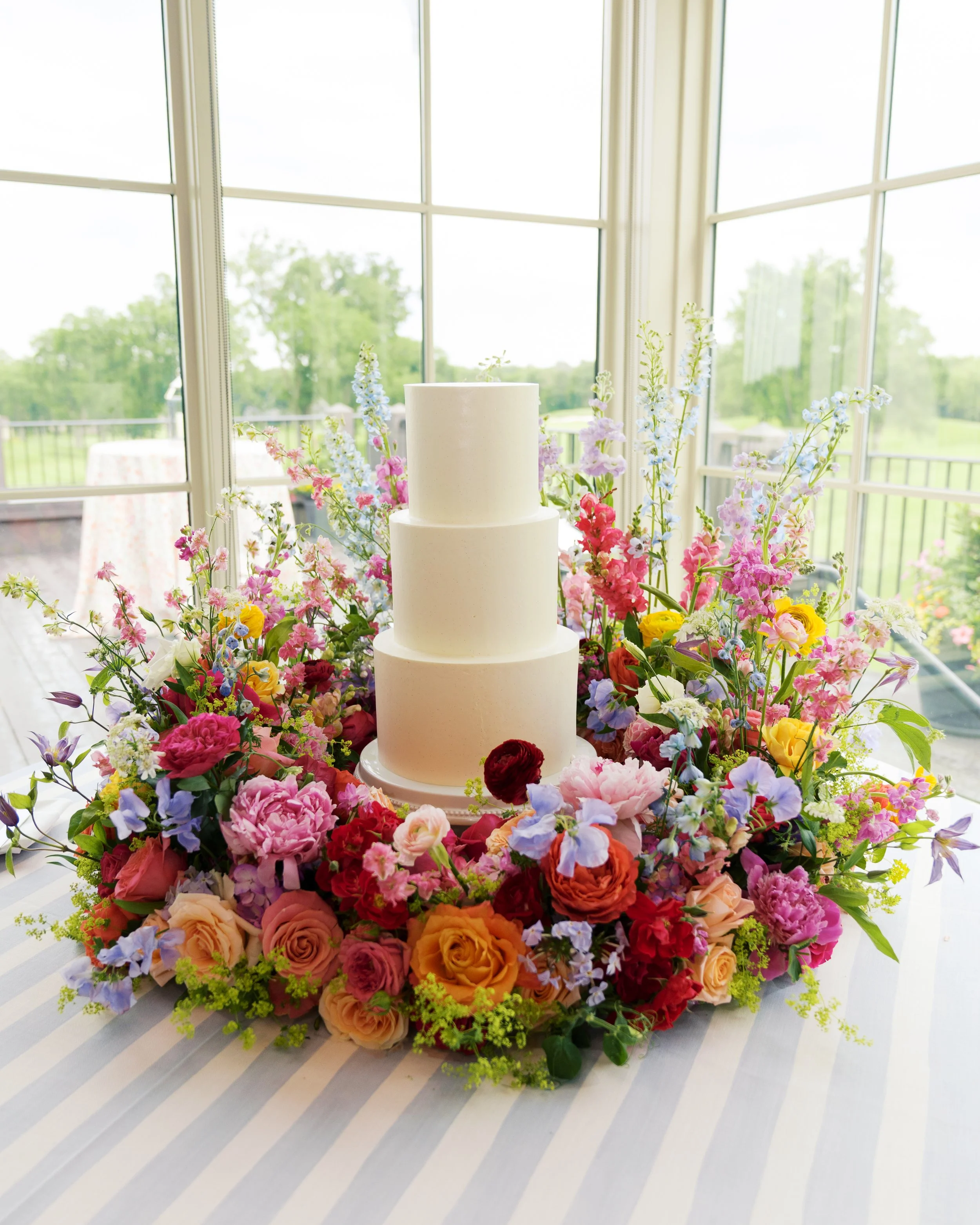 A wedding cake surrounded by a colorful flower arrangement on a table near large windows with a scenic outdoor view.