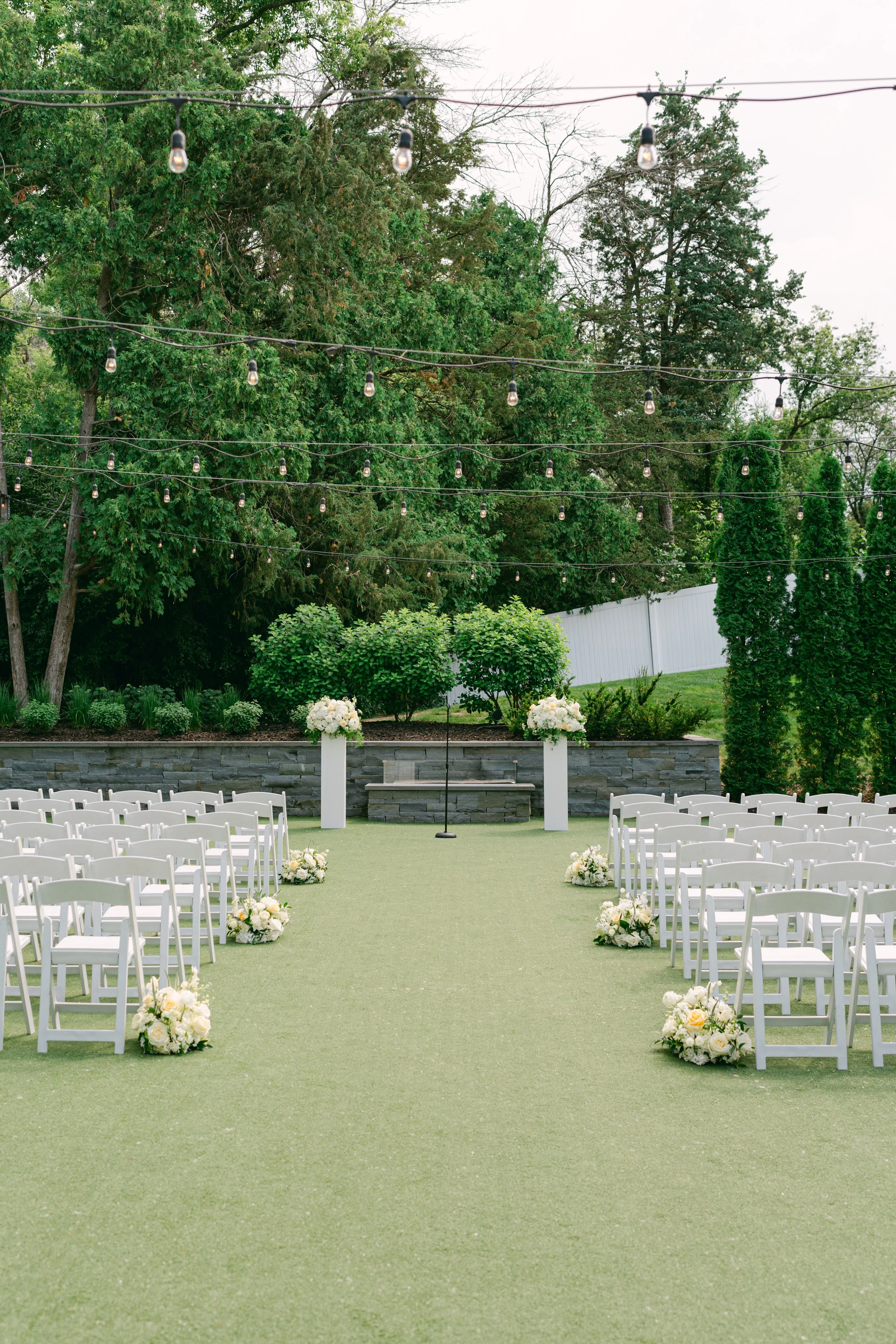 Outdoor wedding ceremony setup with white chairs arranged on a green carpet aisle, floral arrangements at the start of the aisle, and a backdrop of trees and a white fence, with string lights overhead.