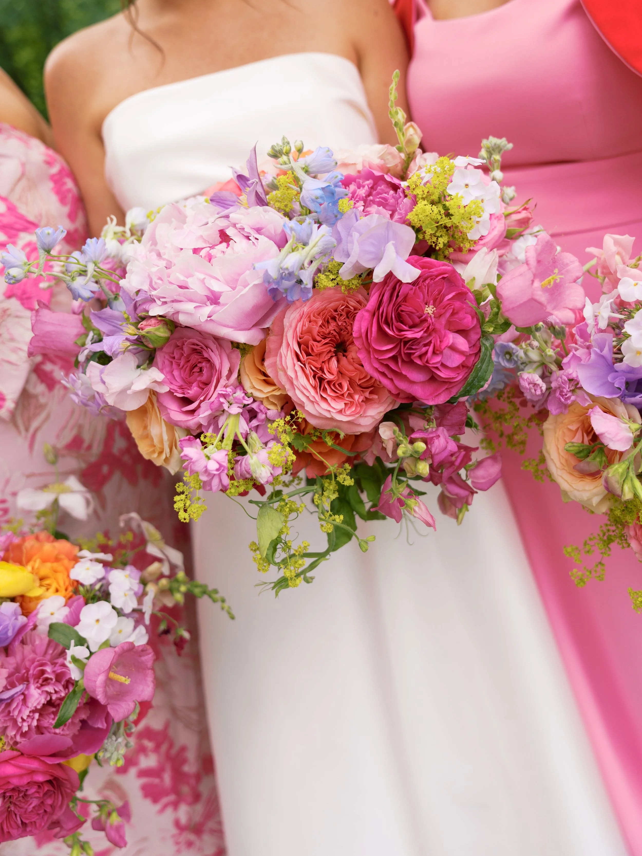 Close-up of women dressed in white and pink dresses holding a large bouquet of pink, purple, yellow, and white flowers.