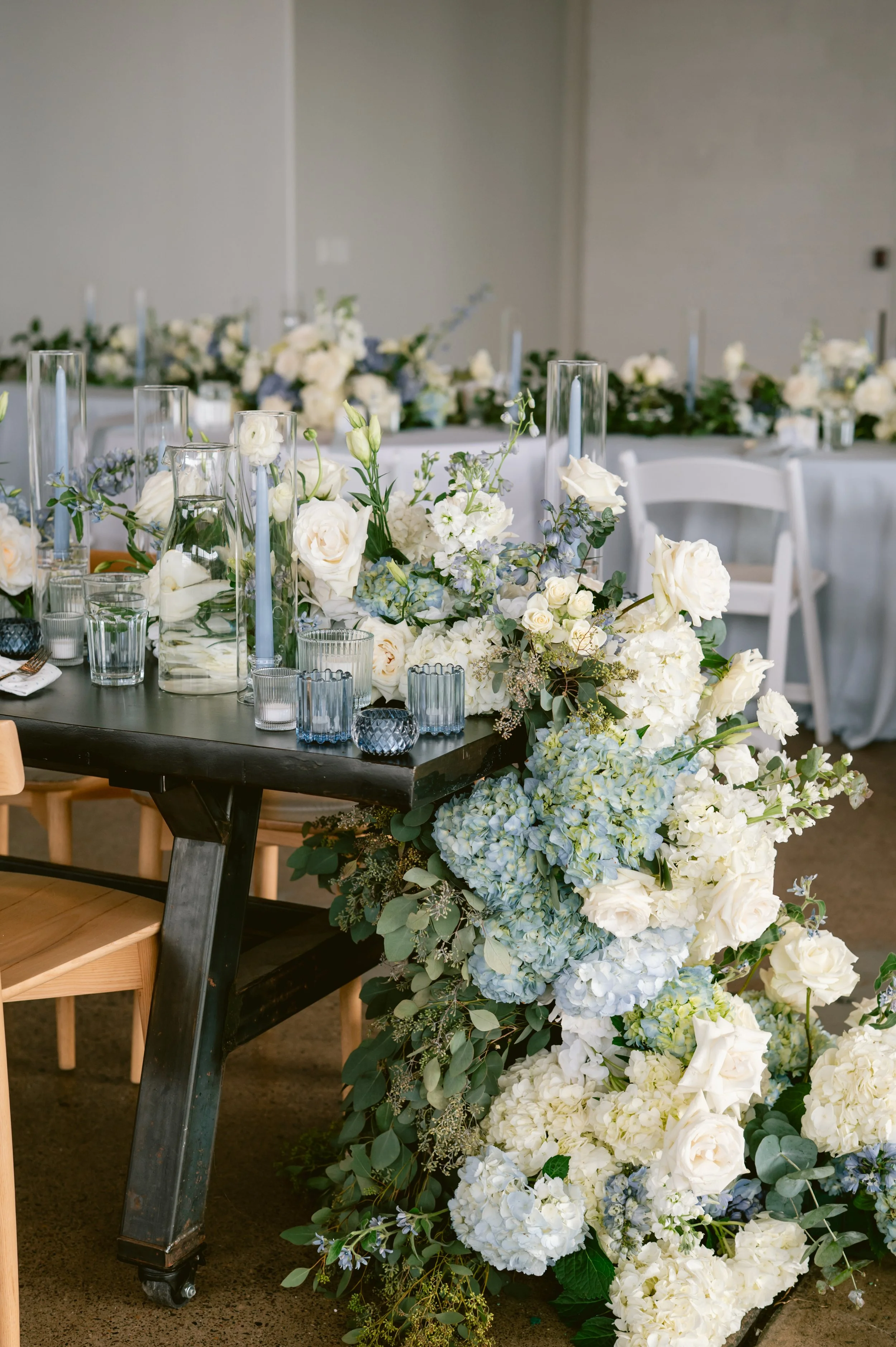 Wedding table decorated with white roses, hydrangeas, greenery, and tall candles in glass holders, with a large floral arrangement cascading from the table to the floor.