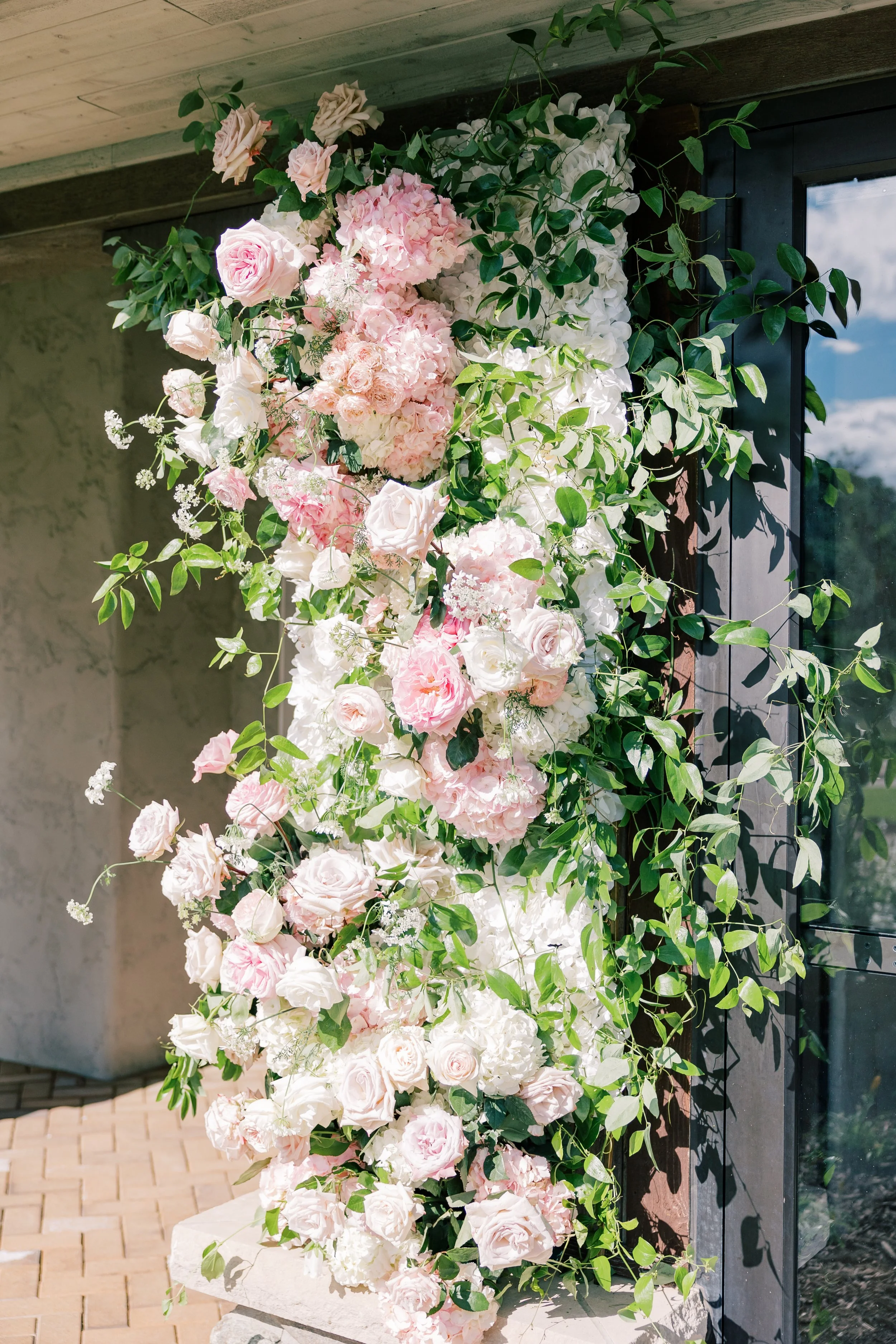 A cascading floral arrangement with pink and white roses, hydrangeas, and green foliage next to a window with a mountain view.