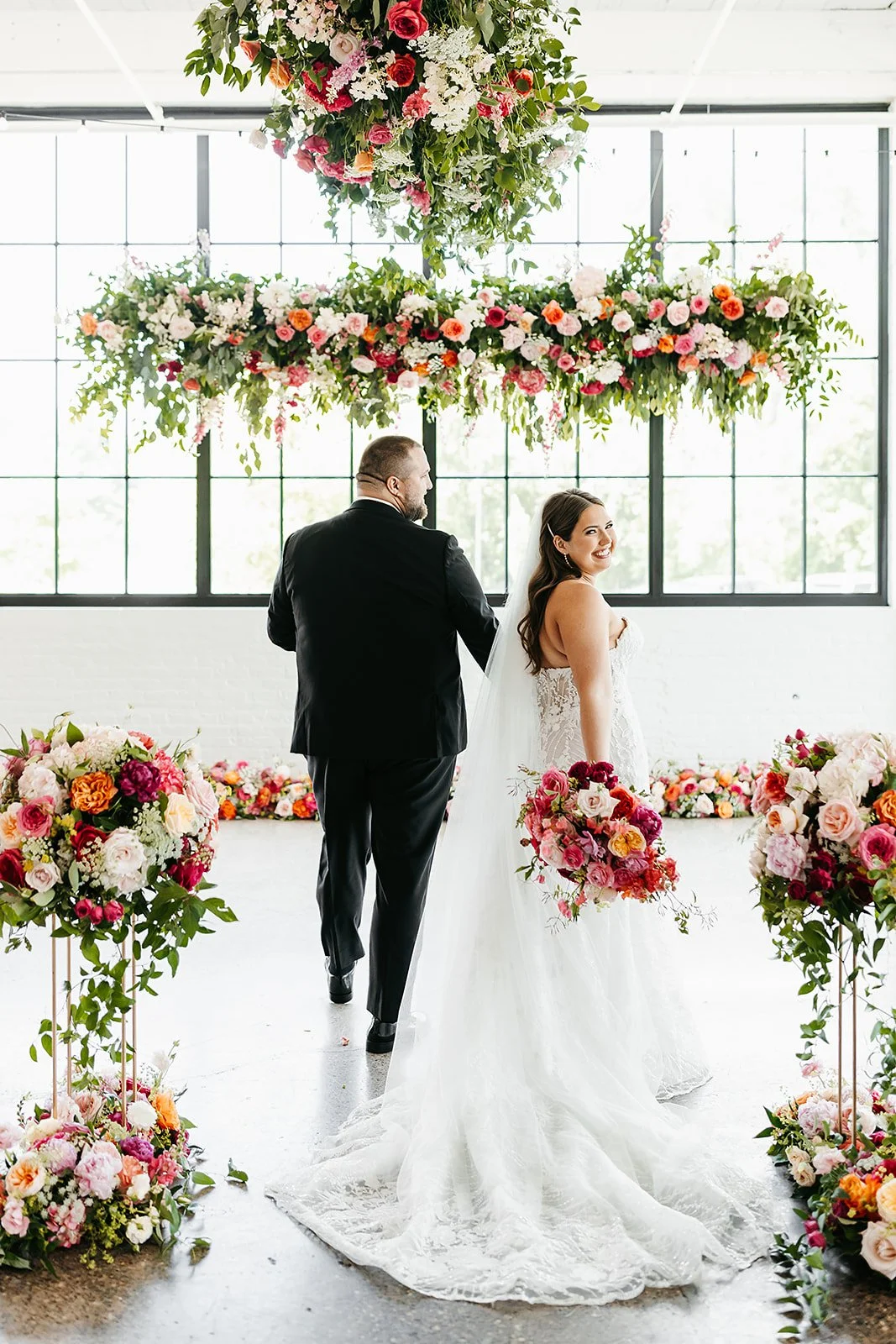 A bride and groom exchanging vows during their wedding ceremony, surrounded by floral arrangements and large hanging floral decorations with large windows in the background.