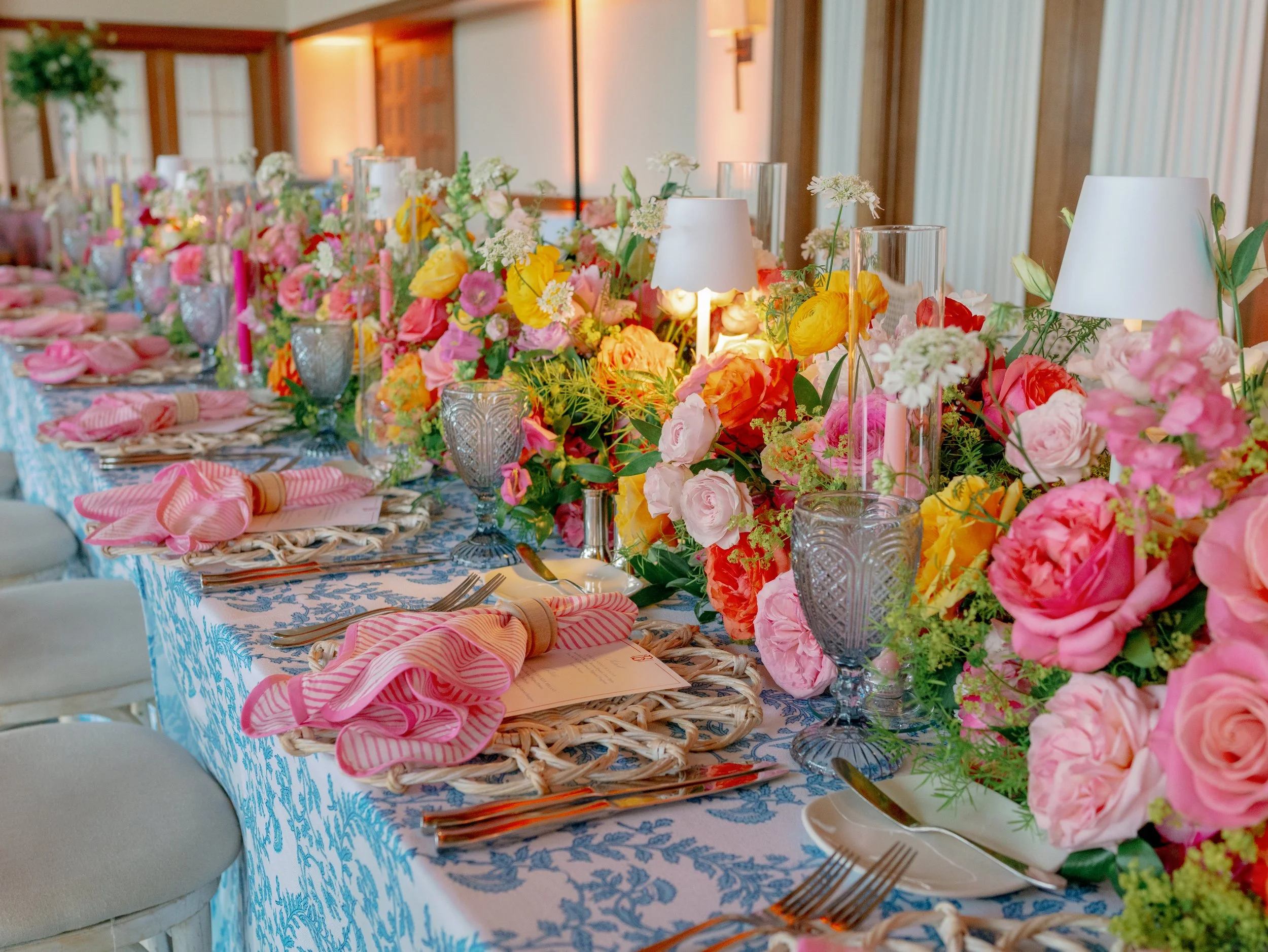 A long banquet table decorated with a blue and white patterned tablecloth, colorful floral centerpieces, pink and white napkins with napkin rings, glassware, and candles. The room has wood paneling and soft lighting.