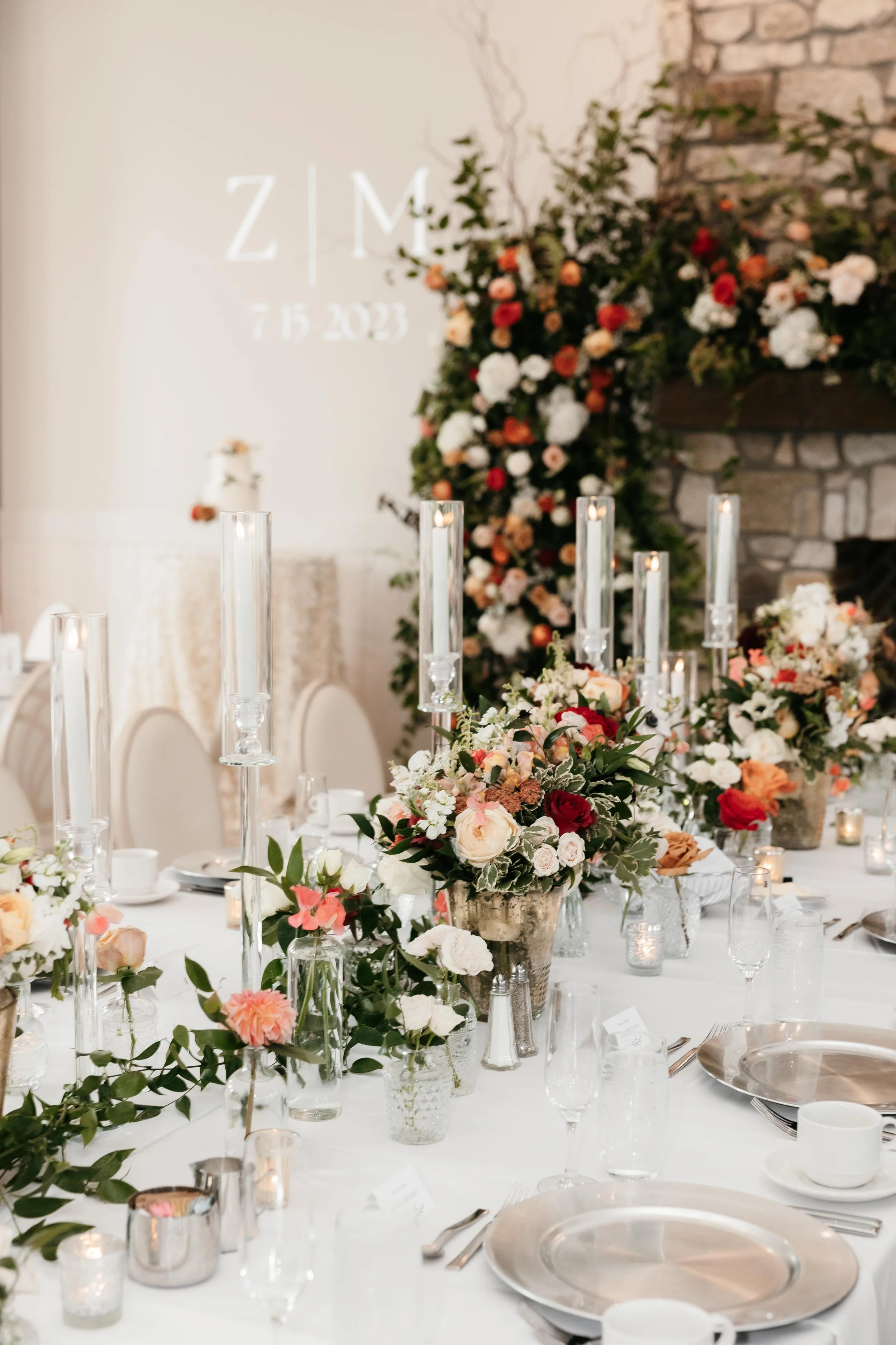 Elegant wedding reception table with floral centerpieces, candles, and place settings. A flower arrangement in gold vases, tall candles in glass holders, and a backdrop with a wall of flowers and a sign that says 'ZIM 7.15.2023'.