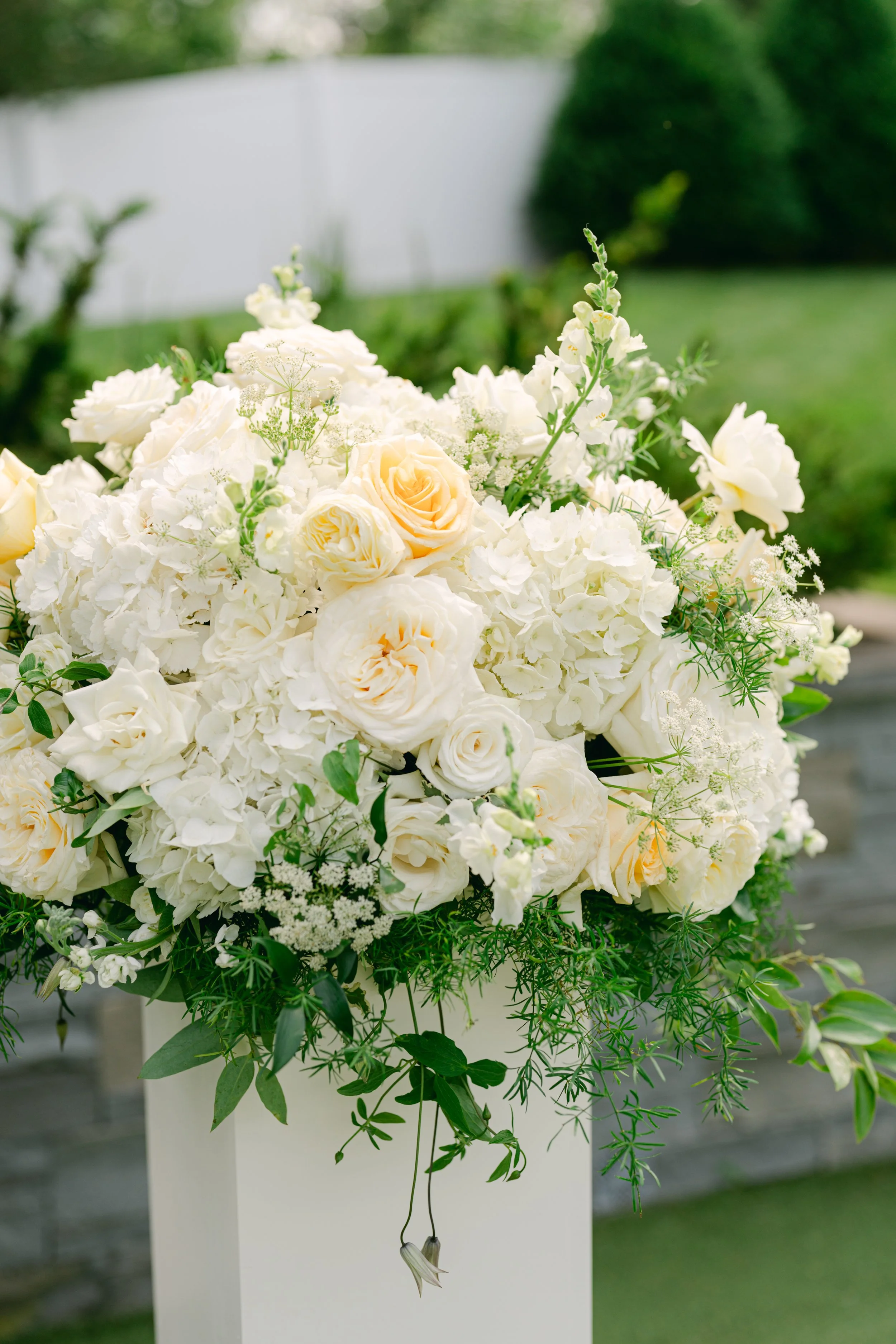A bouquet of white and pale yellow roses and other white flowers with green foliage, placed on a white stand outdoors with a green background.