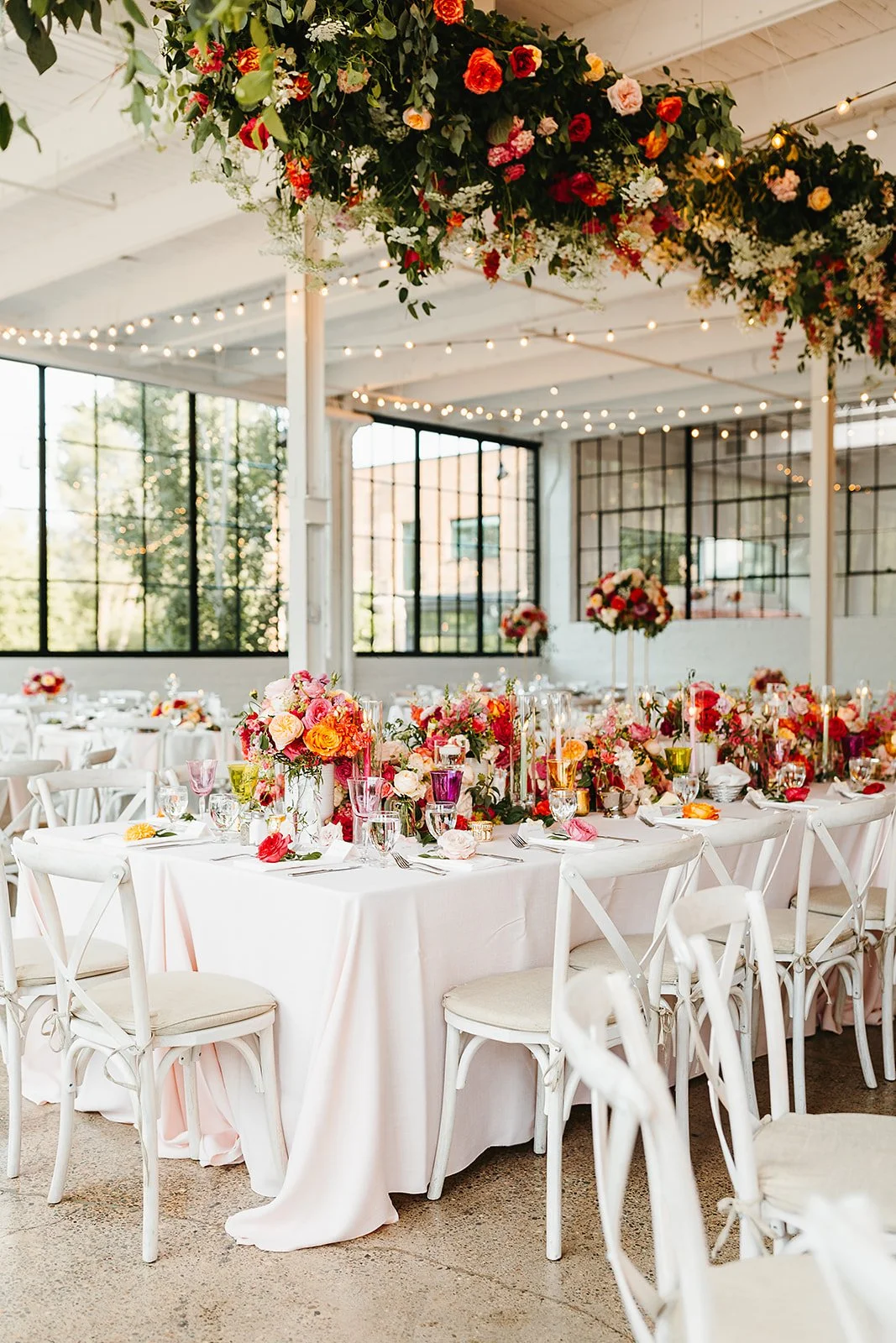 A wedding or celebration reception with white tables and chairs decorated with pink, orange, and white floral centerpieces. Hanging floral arrangement and string lights are overhead inside a bright, industrial-style venue with large windows.