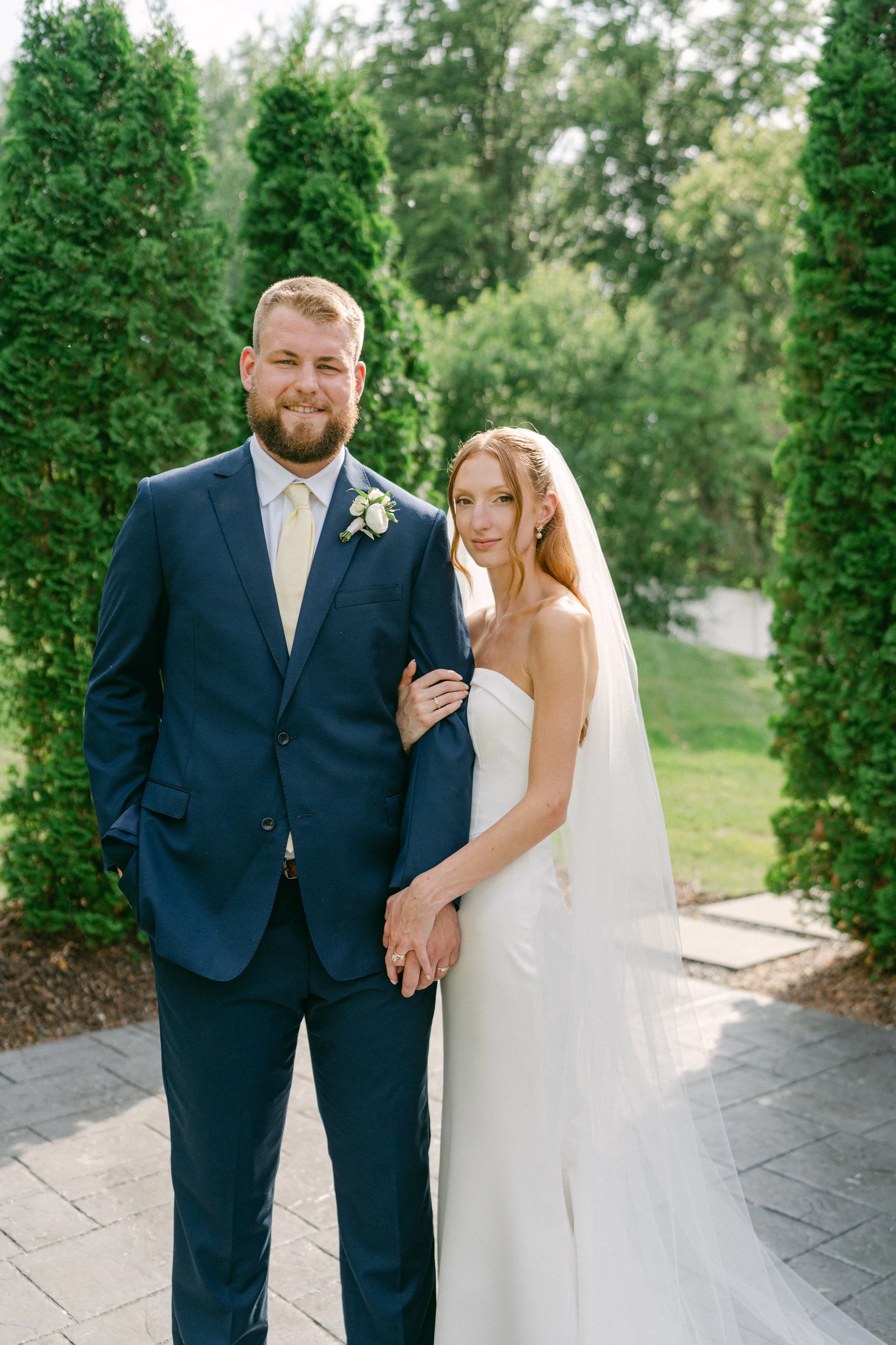 A bride and groom in wedding attire outdoors, surrounded by tall green trees and lush greenery.