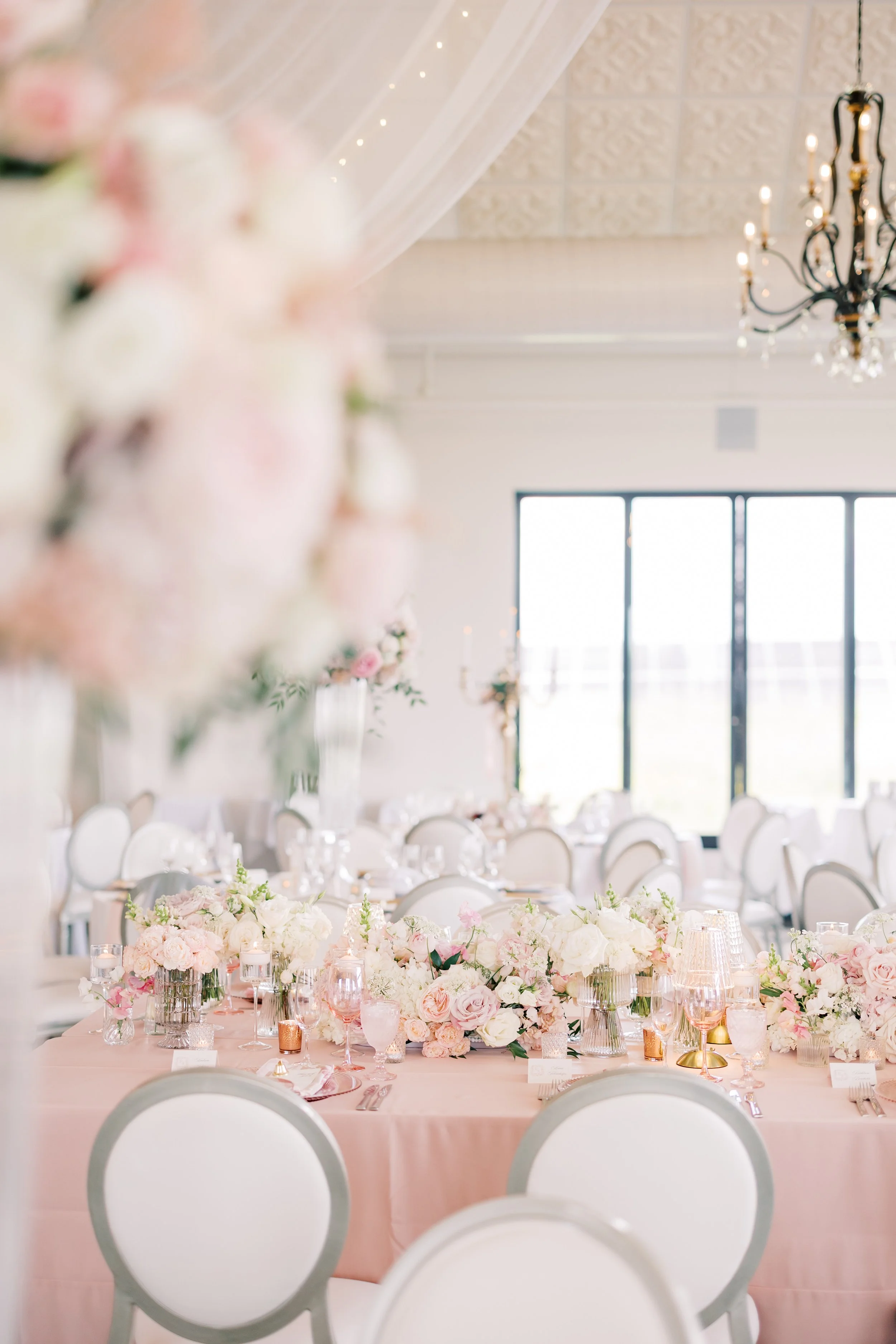 Elegant event table setup with pink and white flowers, gold accents, glassware, and beige chairs in a bright room with large windows and a chandelier.