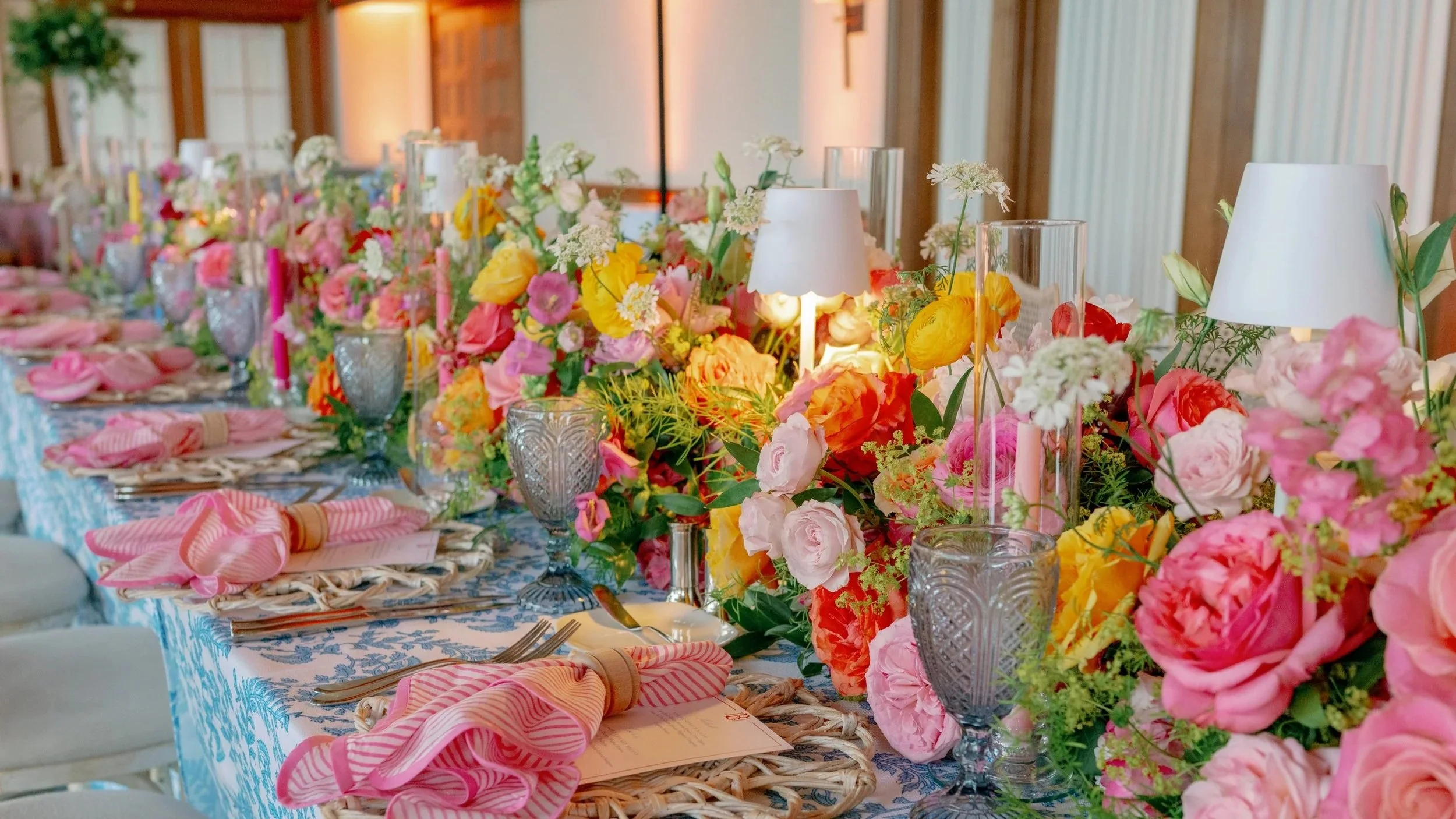 A long banquet table decorated with a colorful floral centerpiece, pink napkins, and elegant glassware and candles, set for a celebration or wedding reception.