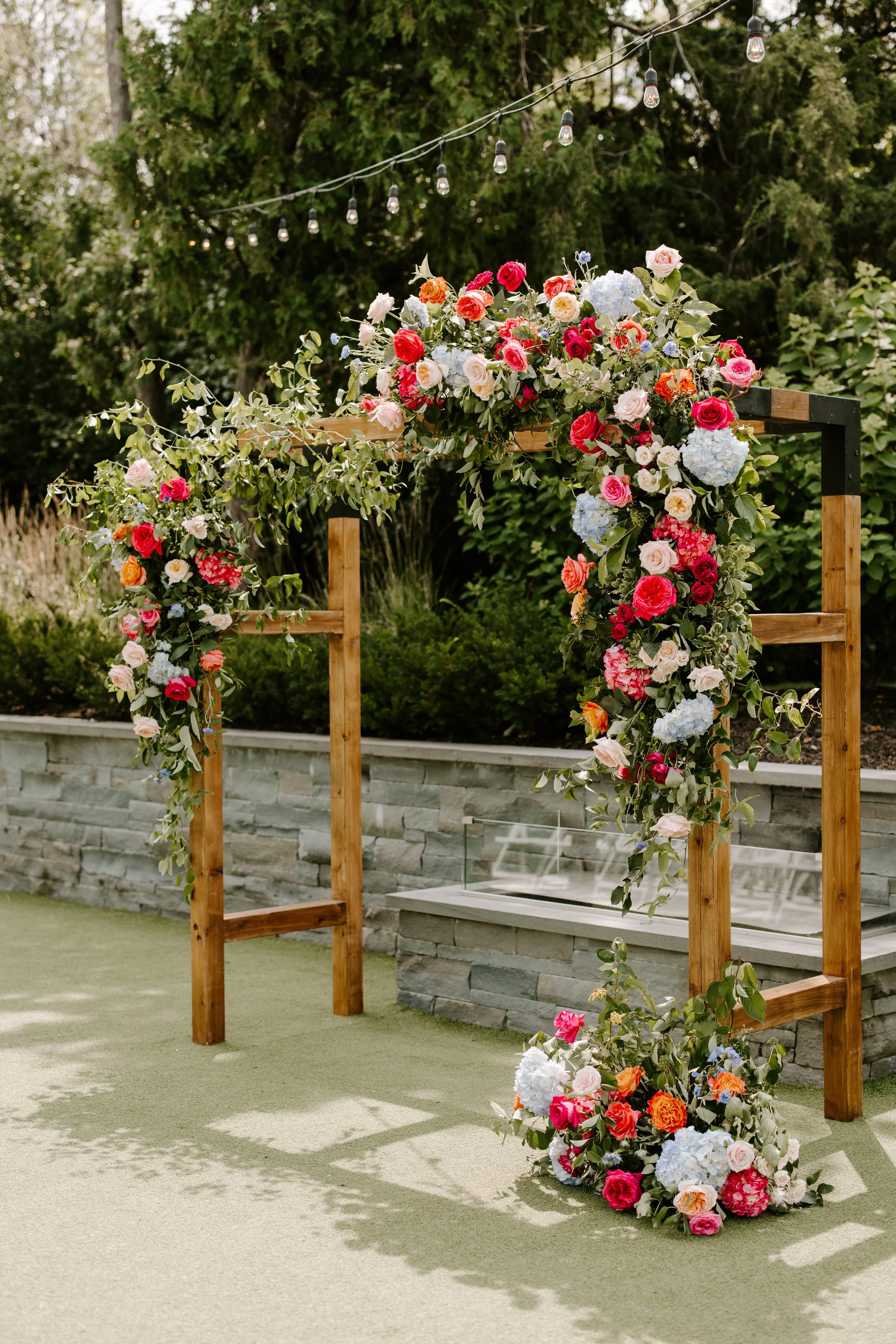 Wooden wedding arch decorated with lush floral arrangements of roses, hydrangeas, and greenery, set outdoors with string lights above and a stone wall in the background.