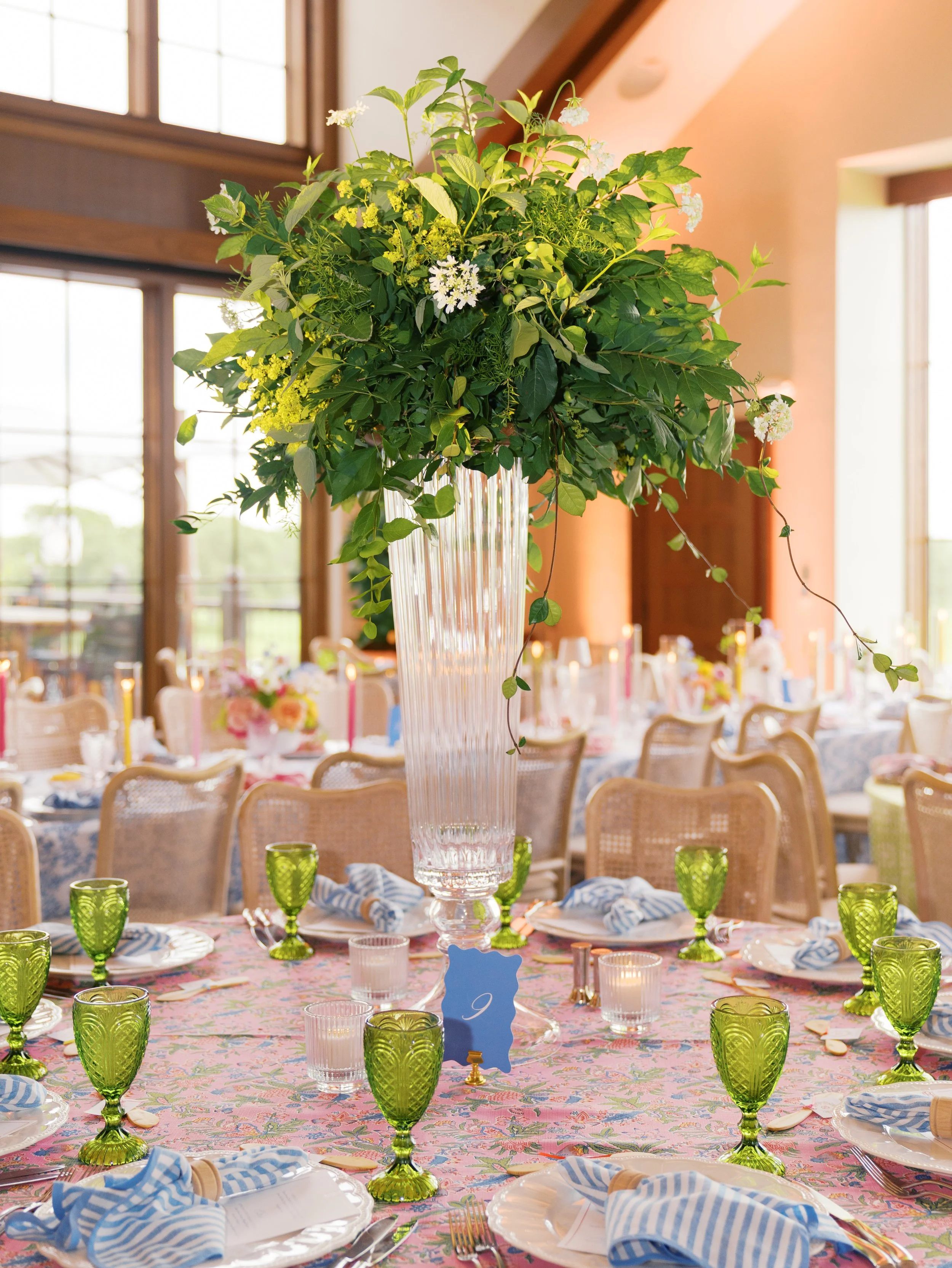 A table decorated for a celebration with green glassware, napkins, and a floral centerpiece in a tall glass vase, set in a bright room with large windows.