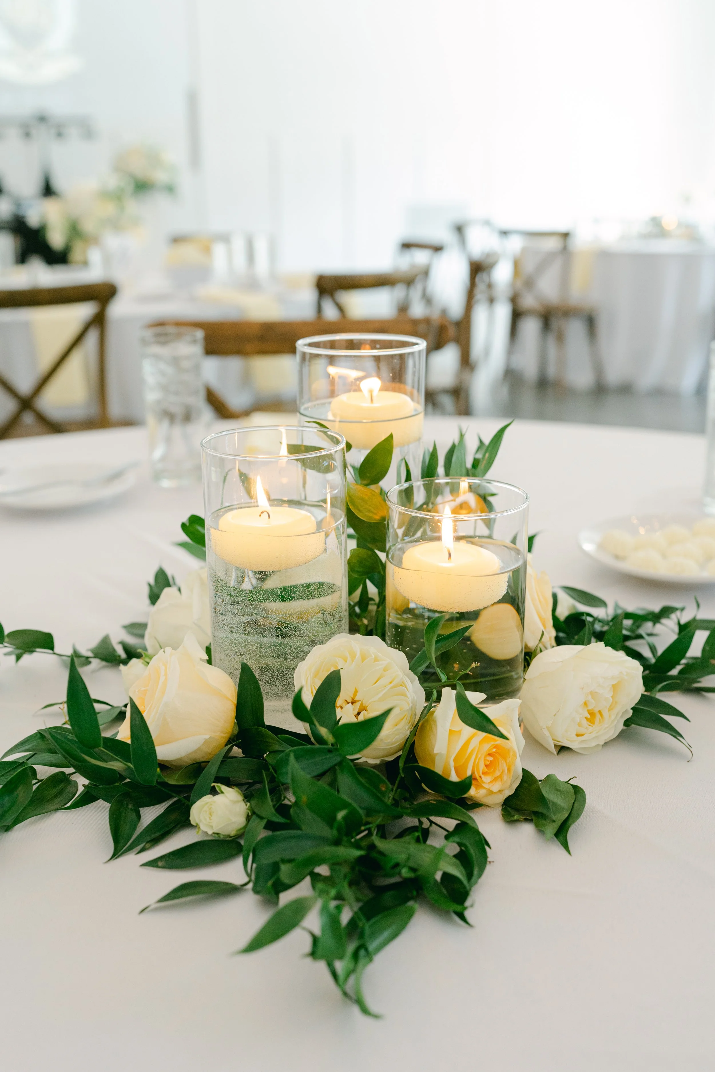 A table centerpiece with floating candles in glass holders surrounded by white roses and green leaves, set in a softly lit event space.