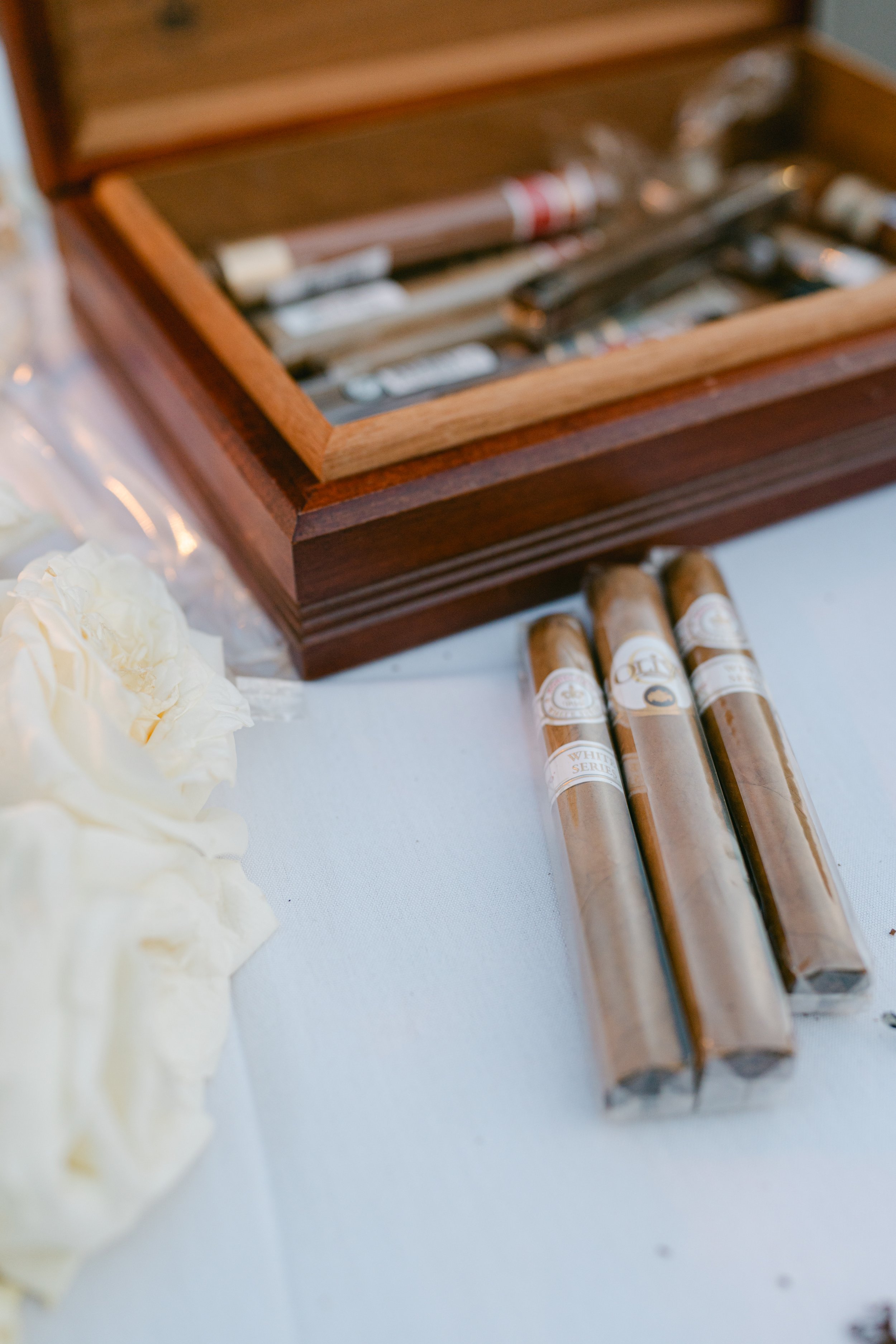 A wooden box containing cigars with labels and a few cigars lying outside the box on a white surface, with some white roses beside it.