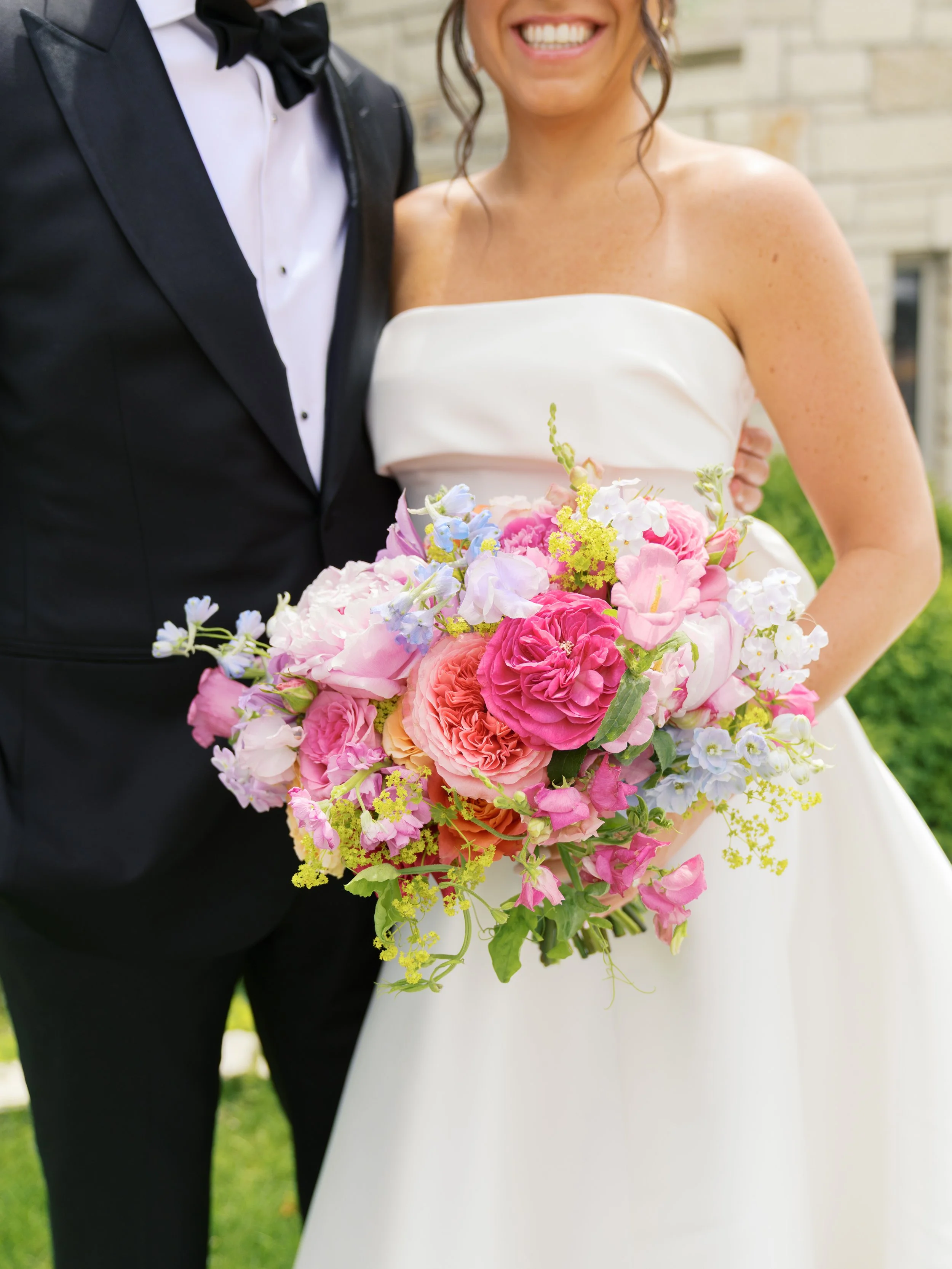 A bride in a white strapless wedding dress holding a colorful bouquet of pink, white, purple, and yellow flowers. A groom in a black tuxedo with a white shirt and black bow tie is standing next to her. They are outdoors in front of a brick building, 
