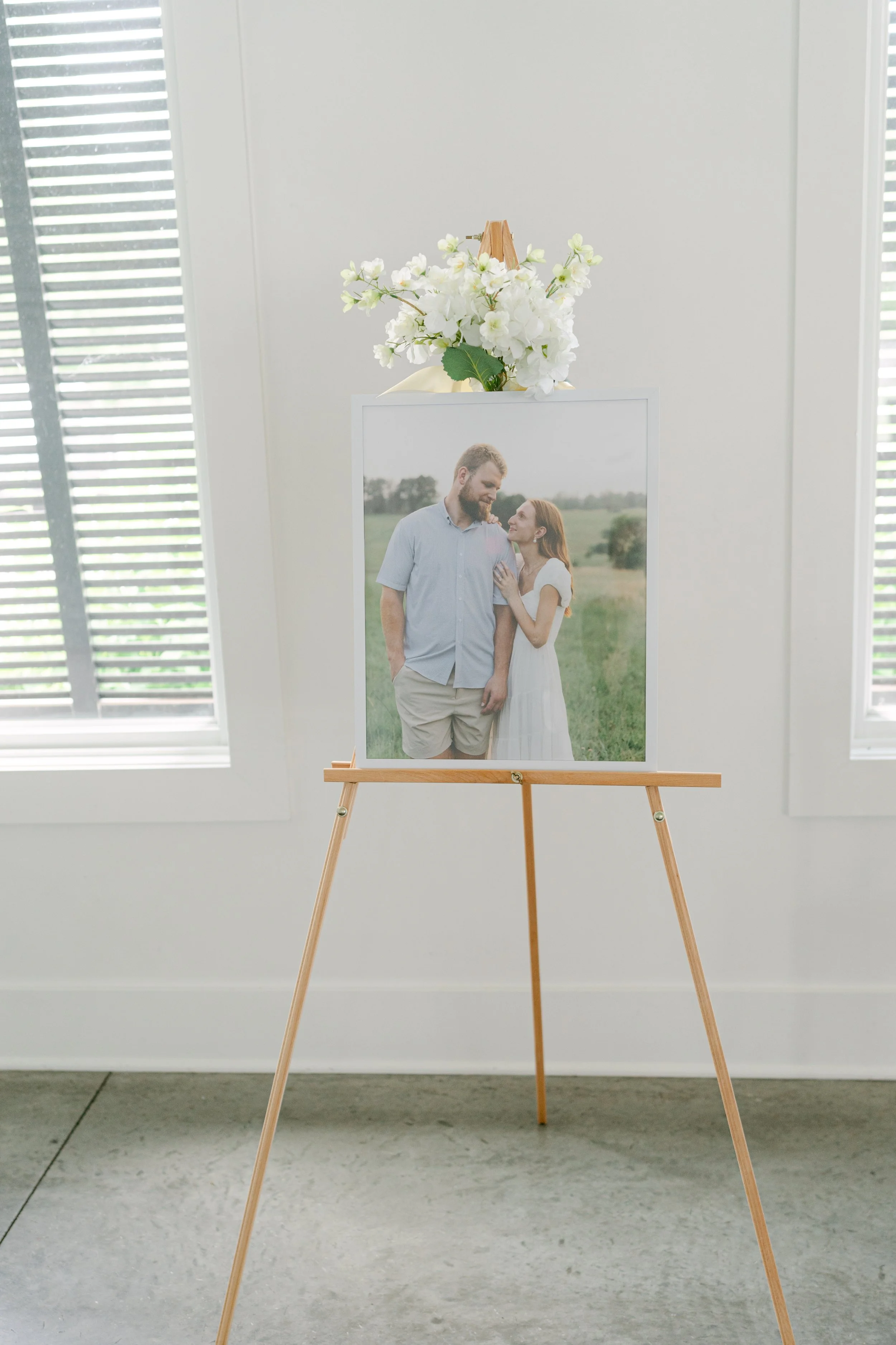 A floral arrangement with white flowers on top of a portrait of a couple standing outdoors, in front of white window blinds.